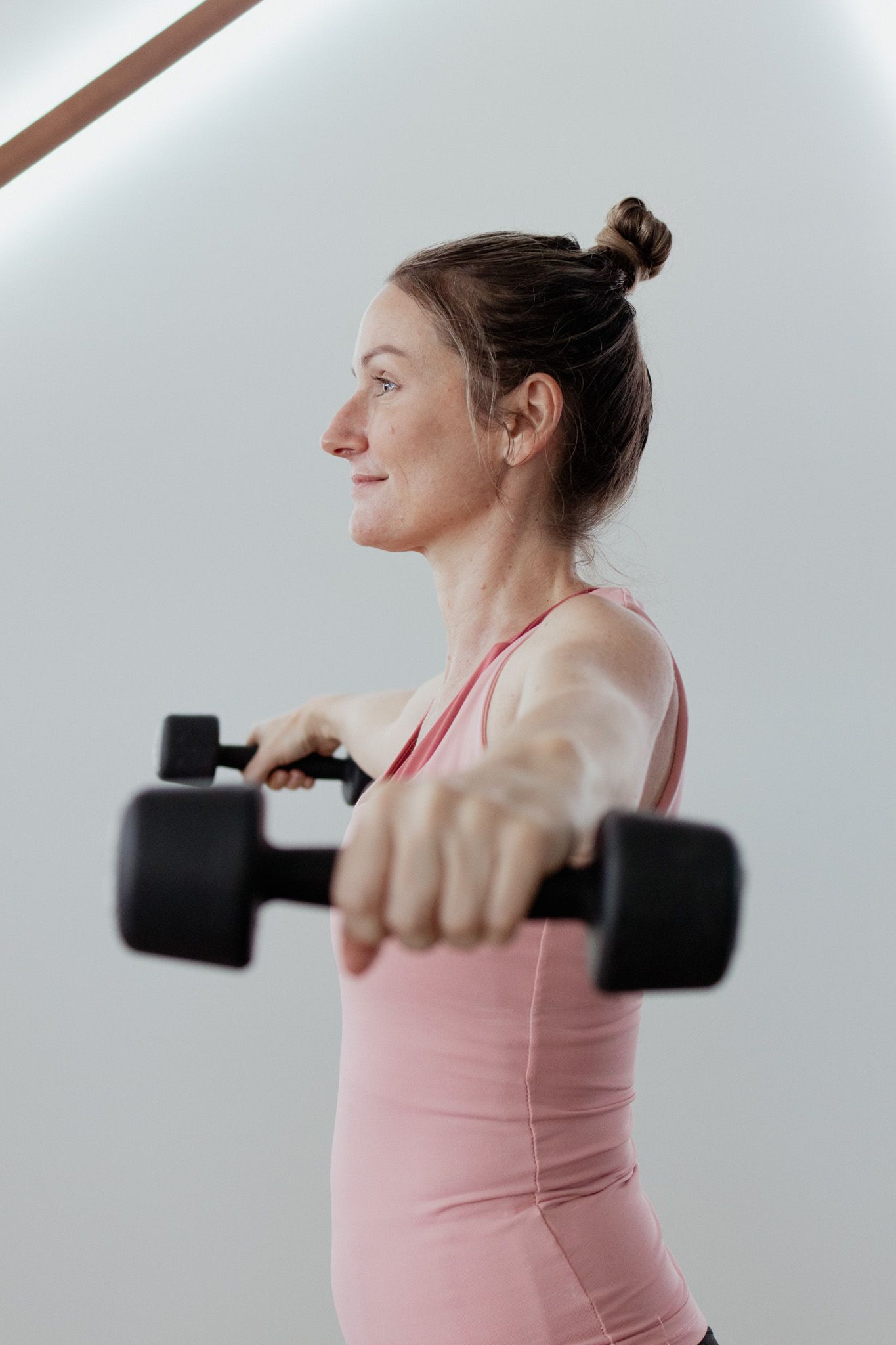 Woman in a pink tank top with her hair in a bun lifts two black dumbbells with arms extended forward during a strength training workout.