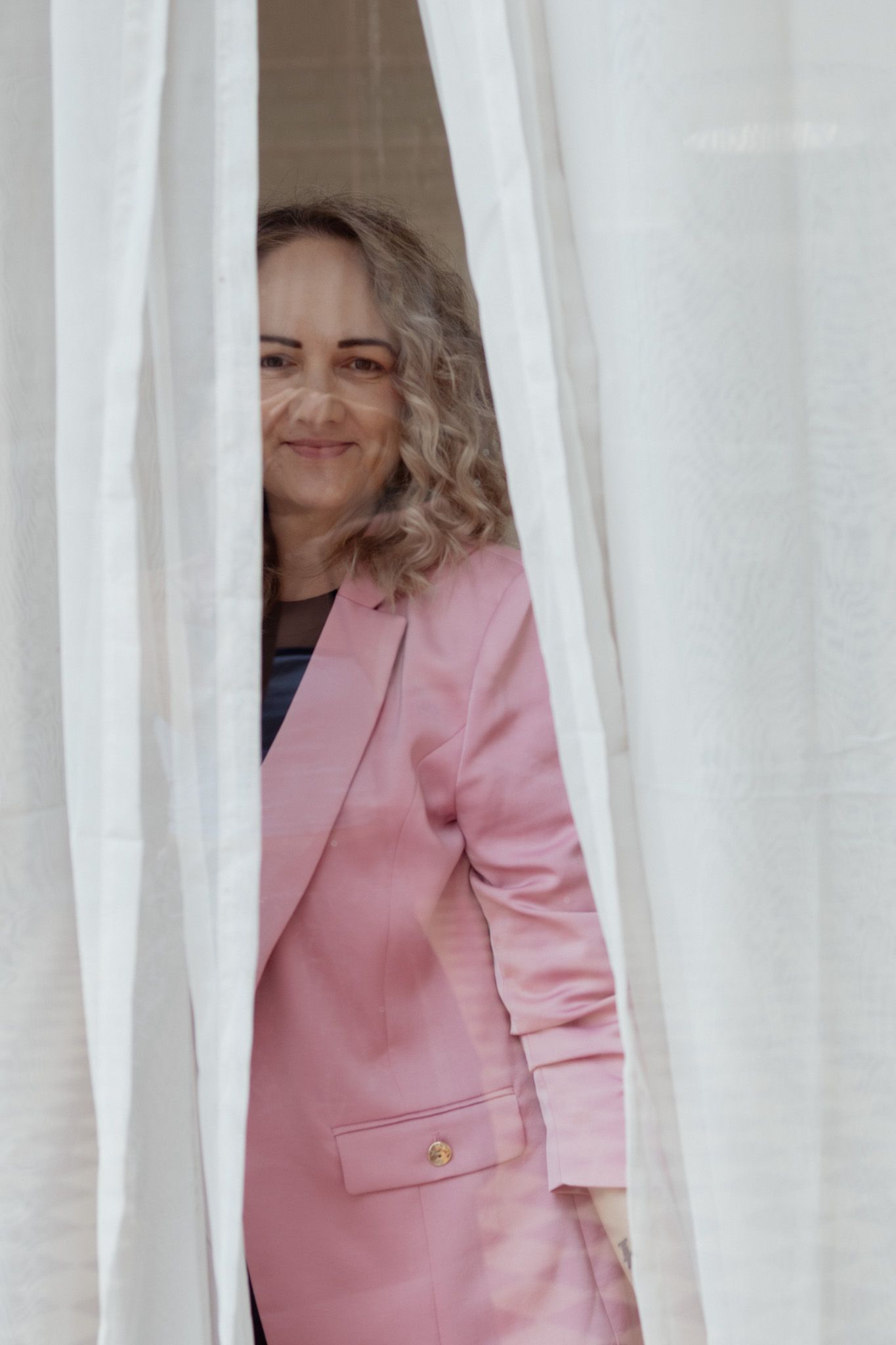 Person in pink blazer peeking through white curtains in sequence.