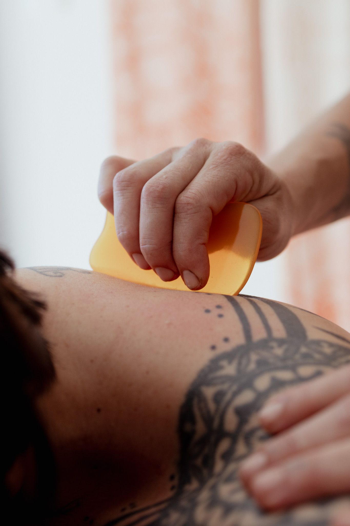 Therapist's hand holding a yellow gua sha tool against a tattooed shoulder during a soothing scraping massage treatment.