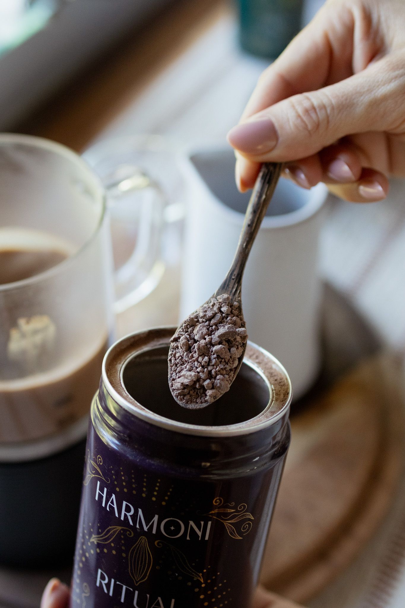 Hand holding a spoon of cacao powder over a purple Harmoni Ritual jar, with a mug of frothy drink in the background.