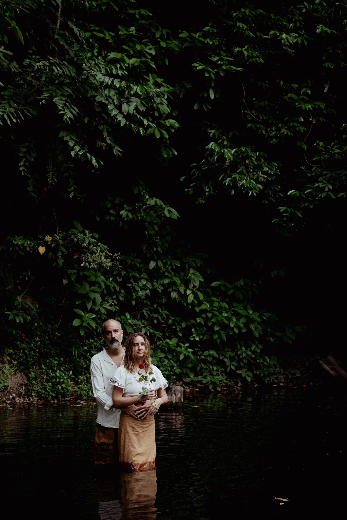 Couple standing together in a wooded area near water in moody lighting.
