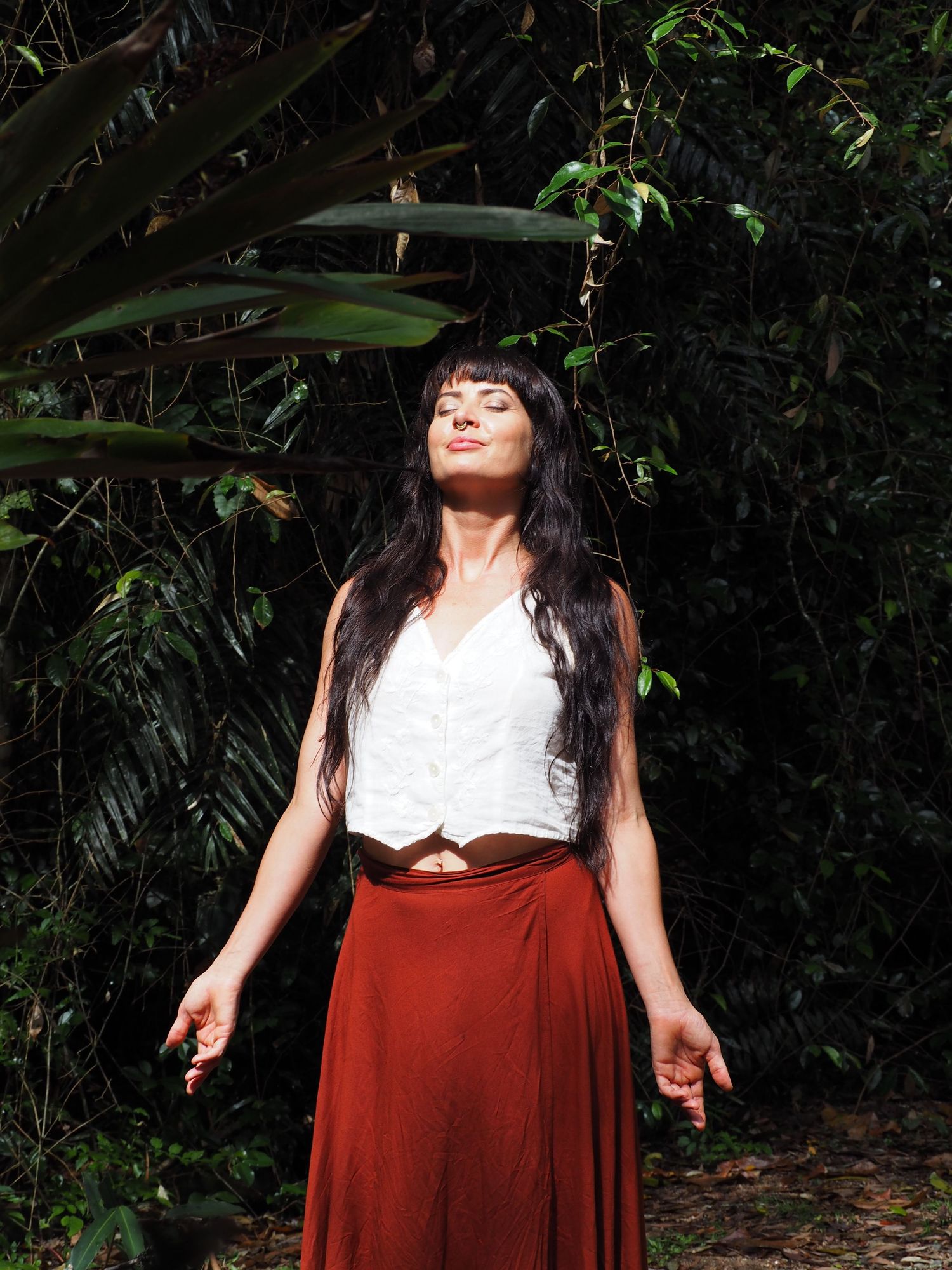 Person in white top and burgundy skirt poses against lush tropical foliage.