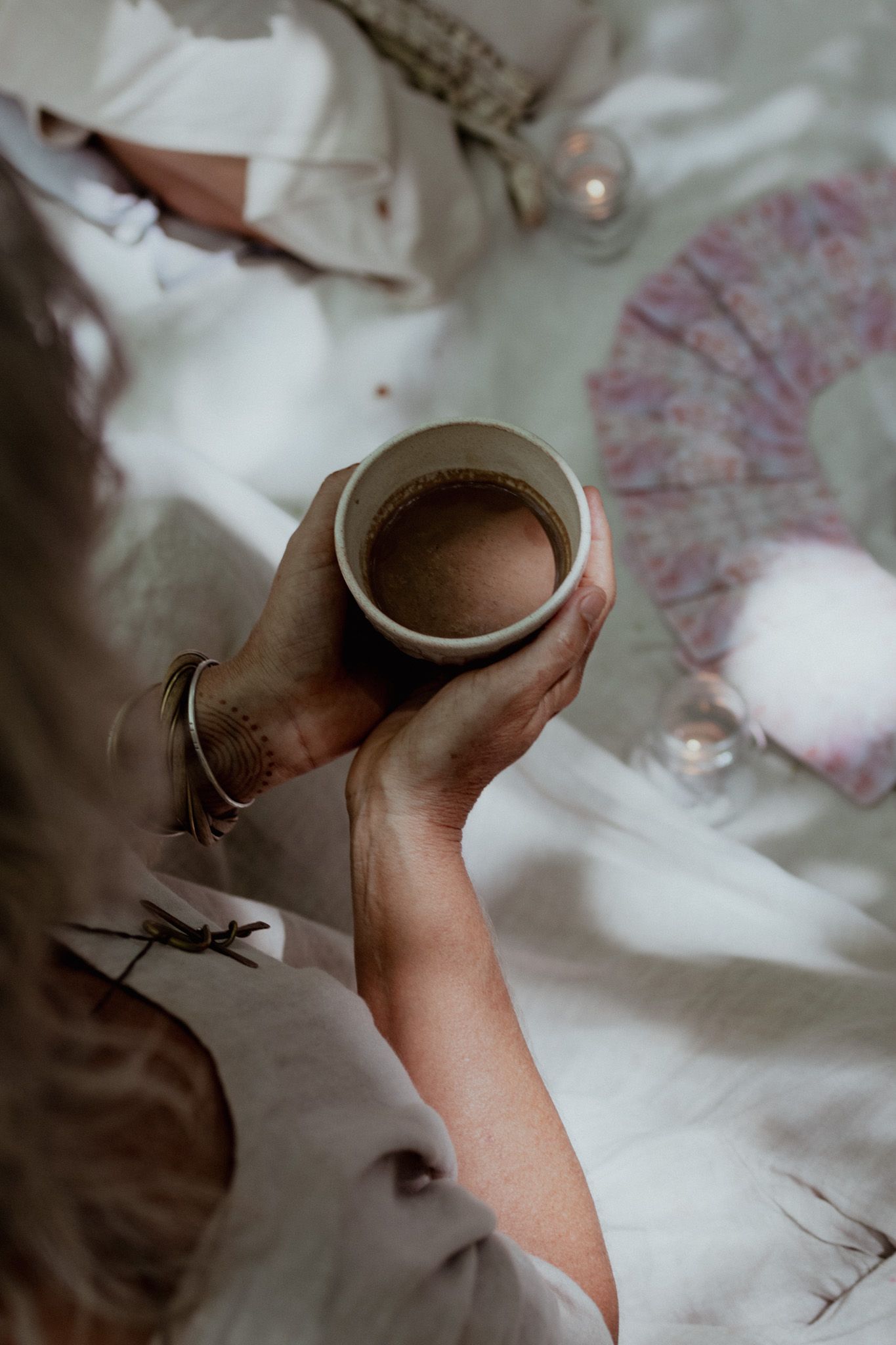 Hands holding a ceramic cup against soft white and pink floral fabric.