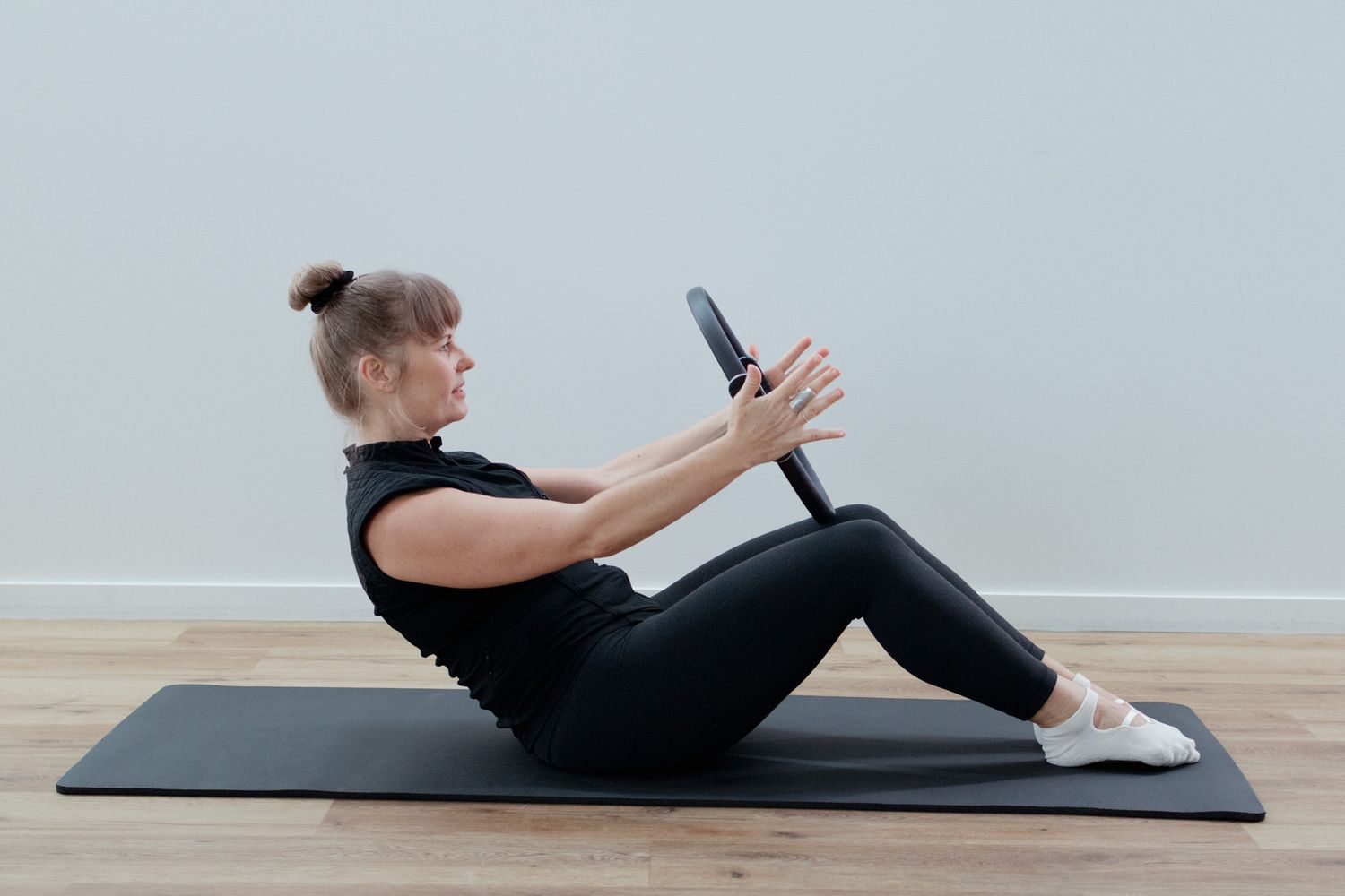 A woman in black activewear sits on a mat performing a Pilates ring exercise on a wooden studio floor.