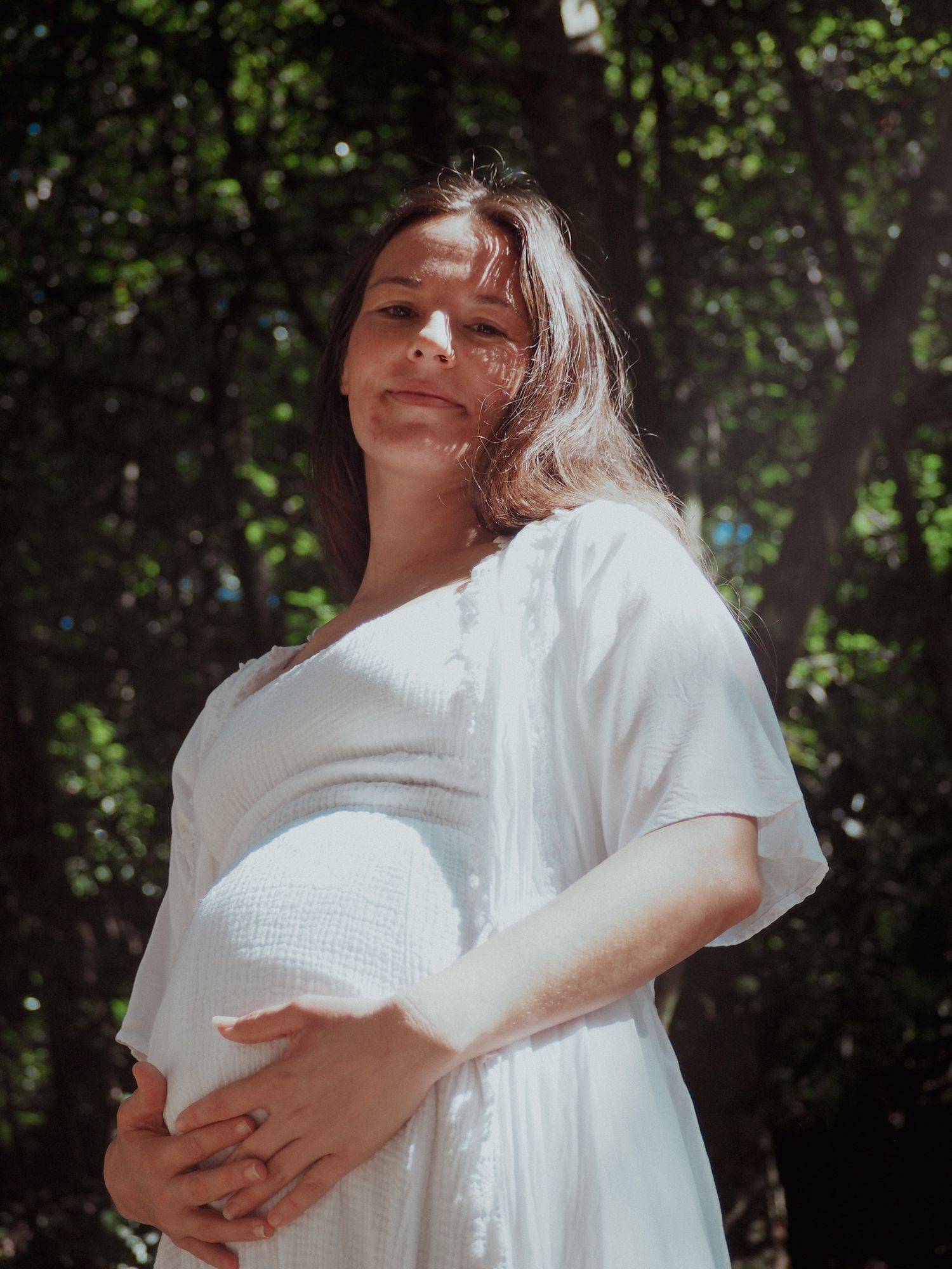 Person in white dress cradling pregnant belly while standing in dappled sunlight outdoors.