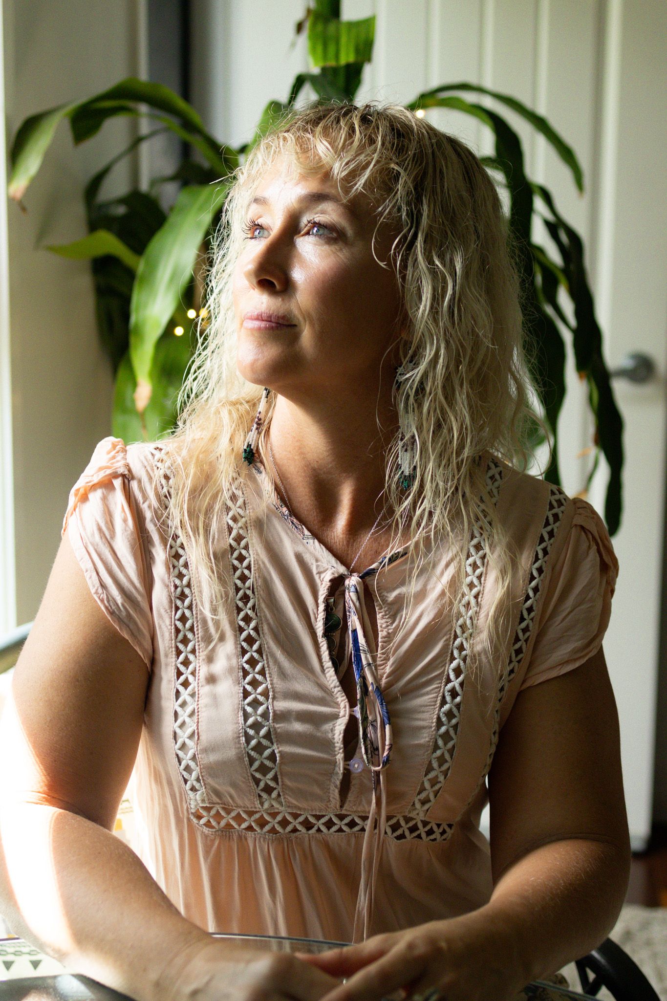 Woman with long wavy blonde hair in a peach lace-trimmed blouse sits by a sunlit window, gazing thoughtfully beside green houseplants.