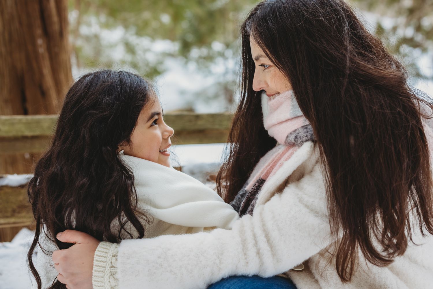 A mother and daughter with dark curly hair share a tender moment embracing in a snowy winter setting.