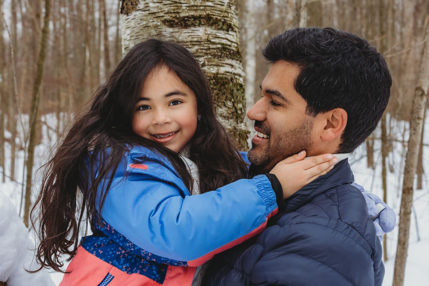 A young girl in a blue jacket smiles while being held by a man in a snowy winter forest.