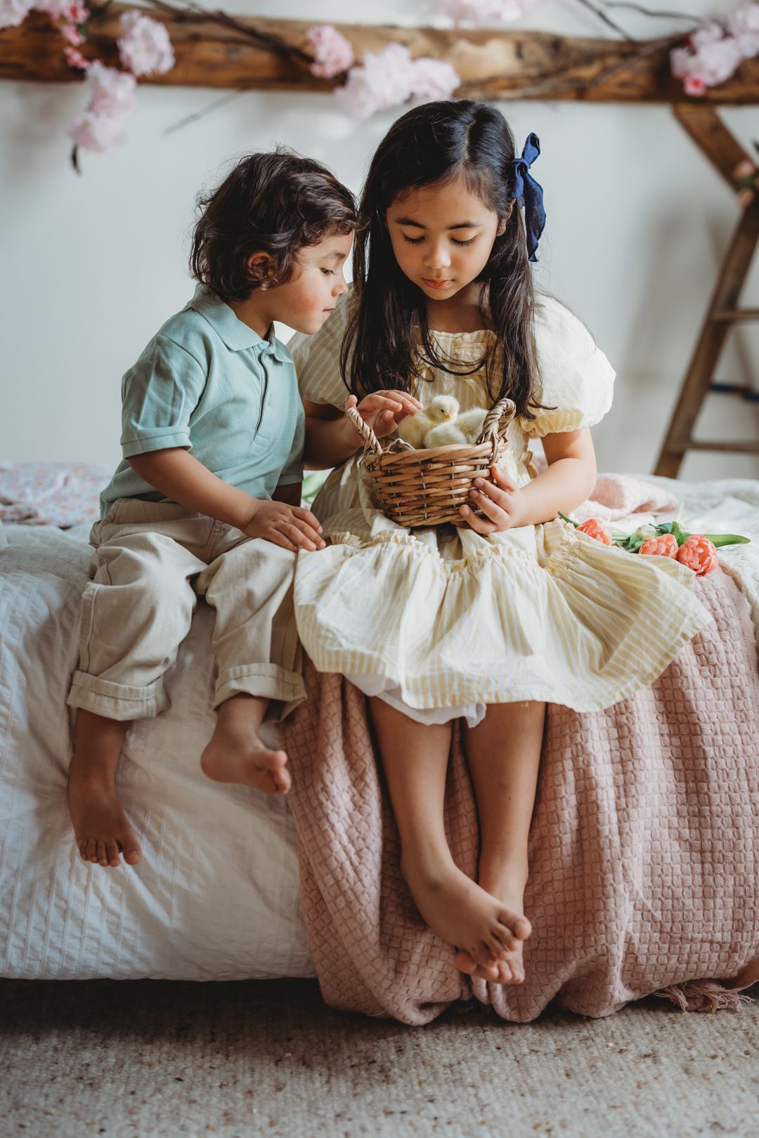 Young girl and boy sitting on bed examining Easter basket with eggs and spring decorations.