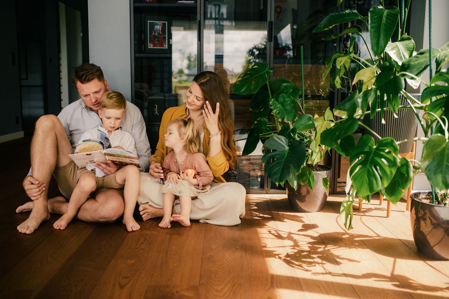 Happy family sitting on sunny hardwood floor reading a colorful book together surrounded by lush indoor plants.