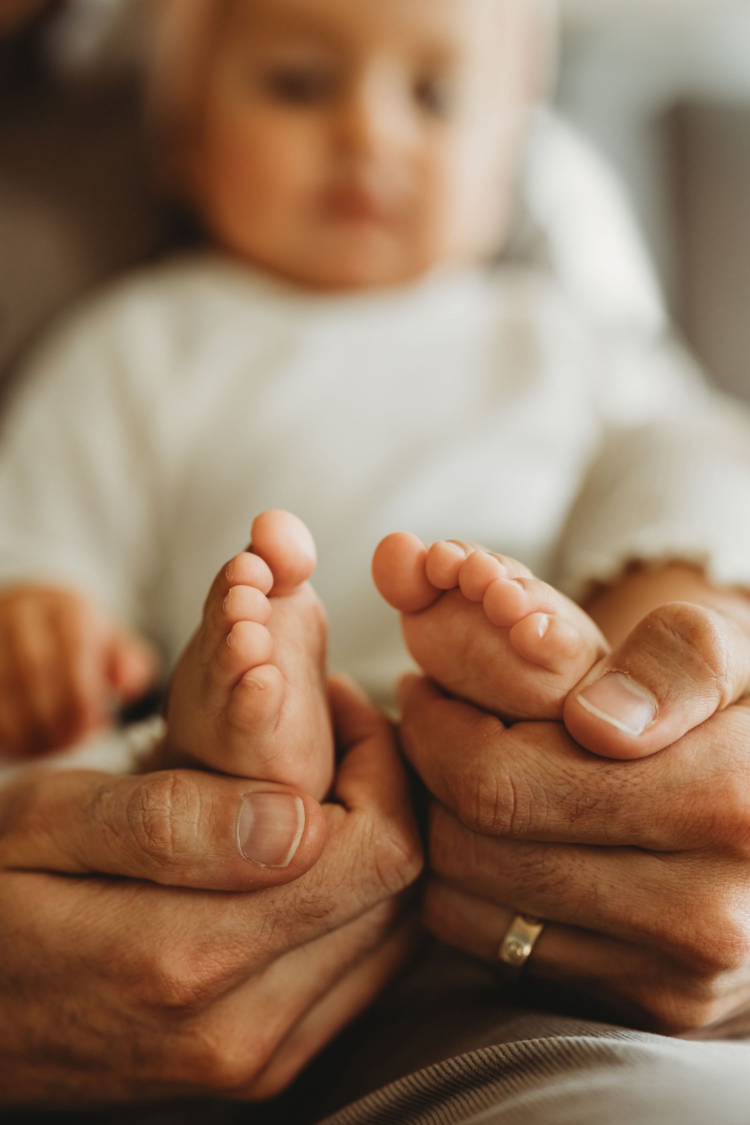 Adult hands gently holding tiny baby feet, with a blurred infant in white outfit in the background.