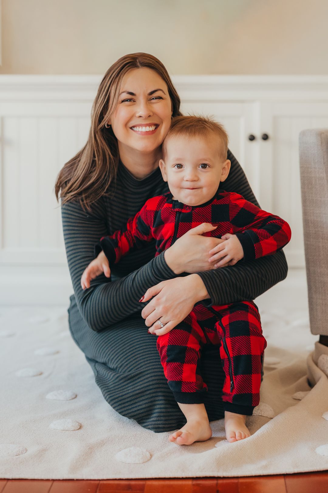 Smiling mother in gray dress hugging baby in red plaid pajamas on white rug indoors.