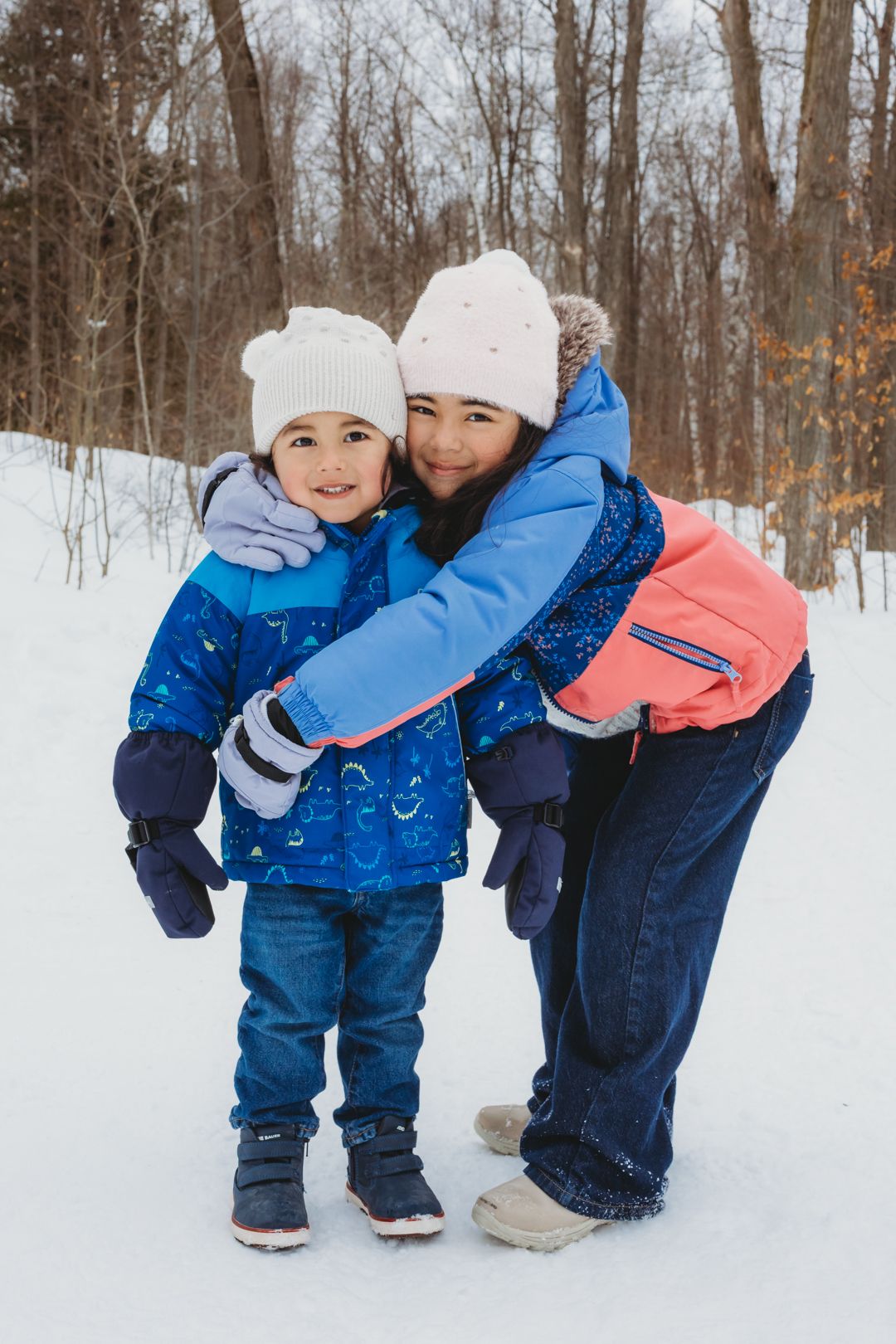 A mother and child in blue winter jackets and white hats embrace outdoors in a snowy forest setting.
