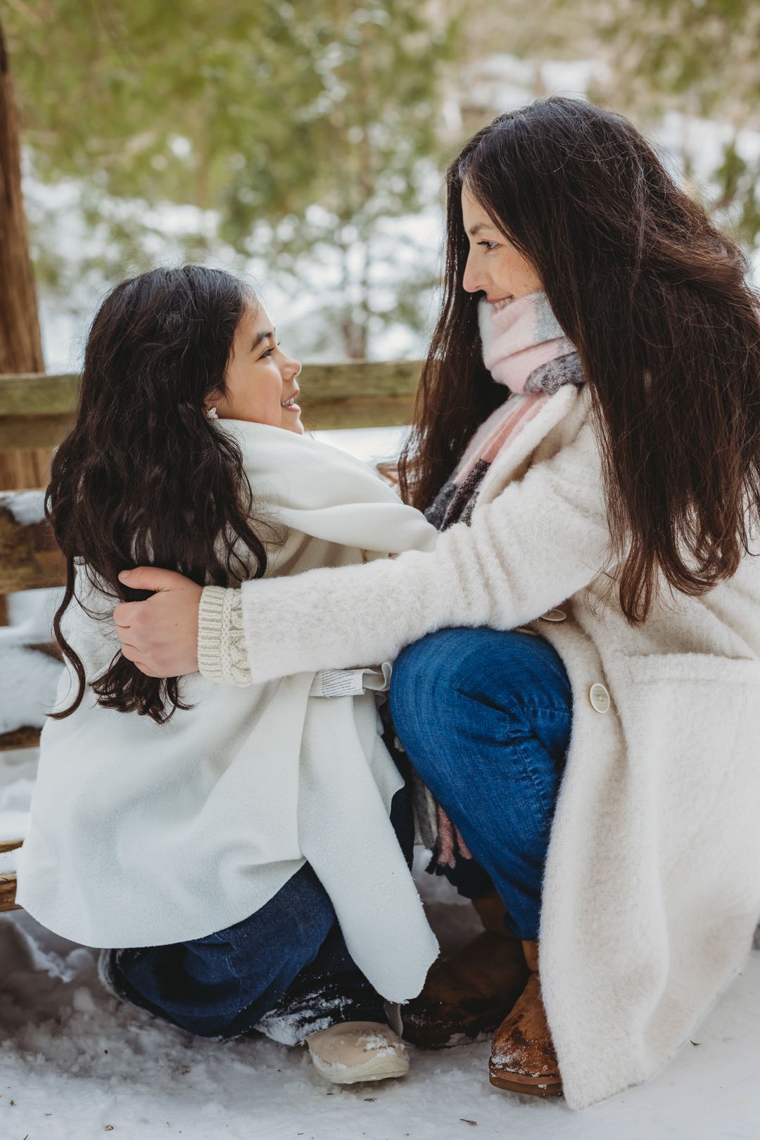 A mother and daughter share a warm embrace outdoors in winter, both wearing white coats and smiling at each other in the snow.