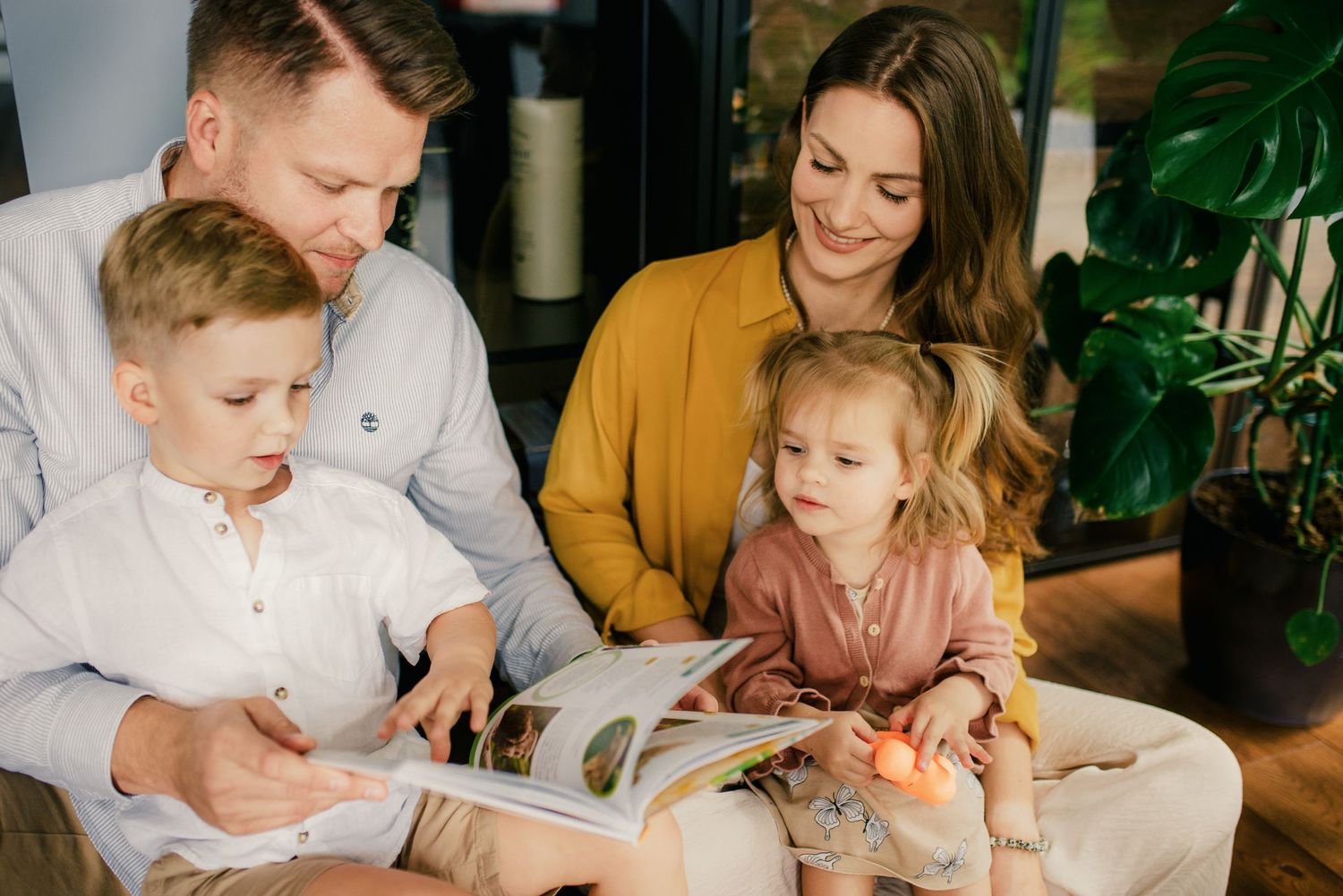 Family of four reading a colorful book together on a cozy sofa at home.
