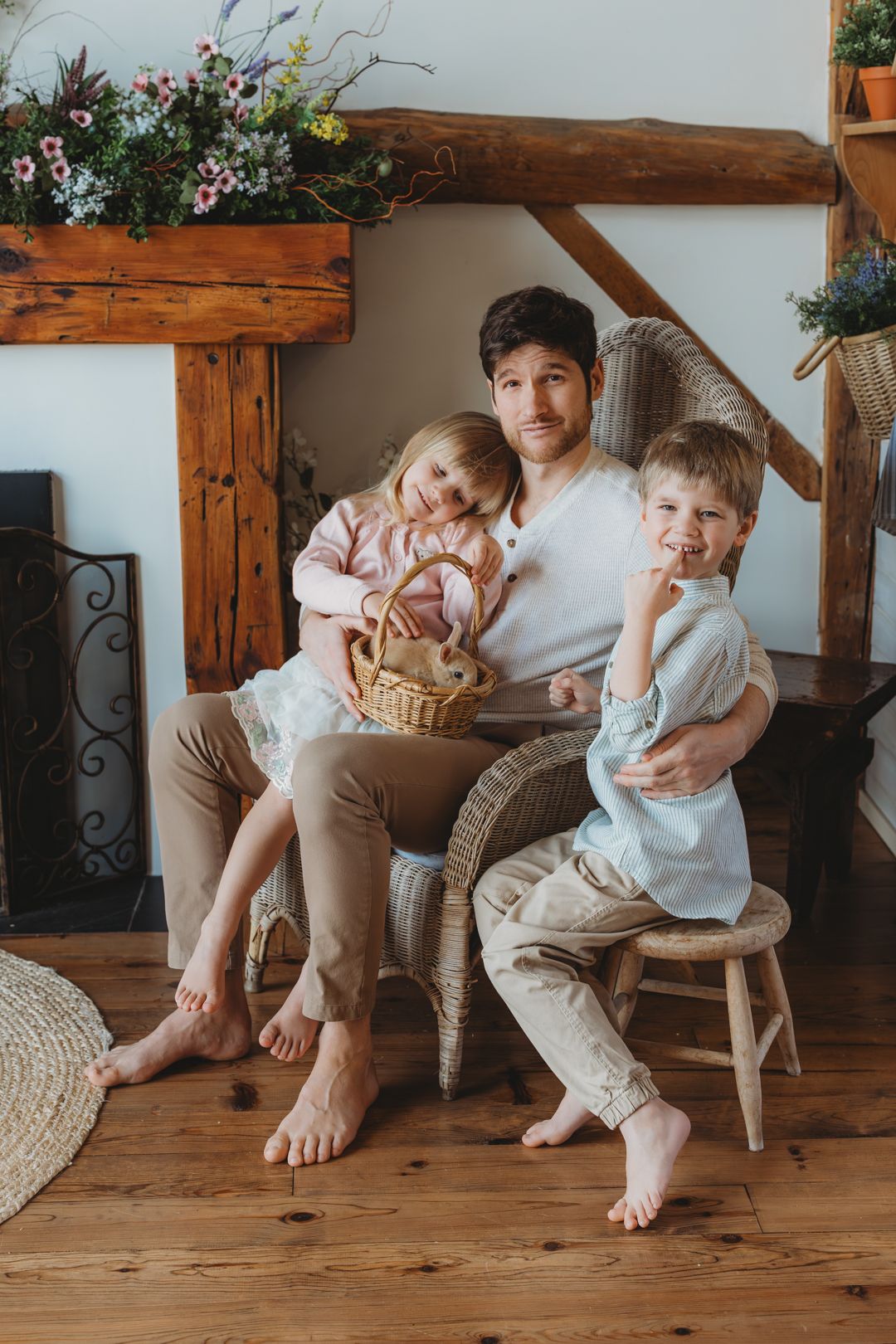 Father in white shirt sits in wicker chair with two young children in a rustic floral interior.