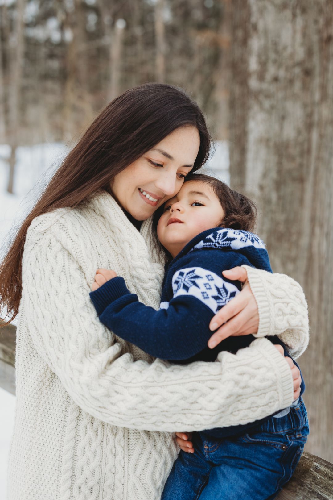 Loving mother hugging her young son in a blue knit sweater outdoors in a snowy winter setting.
