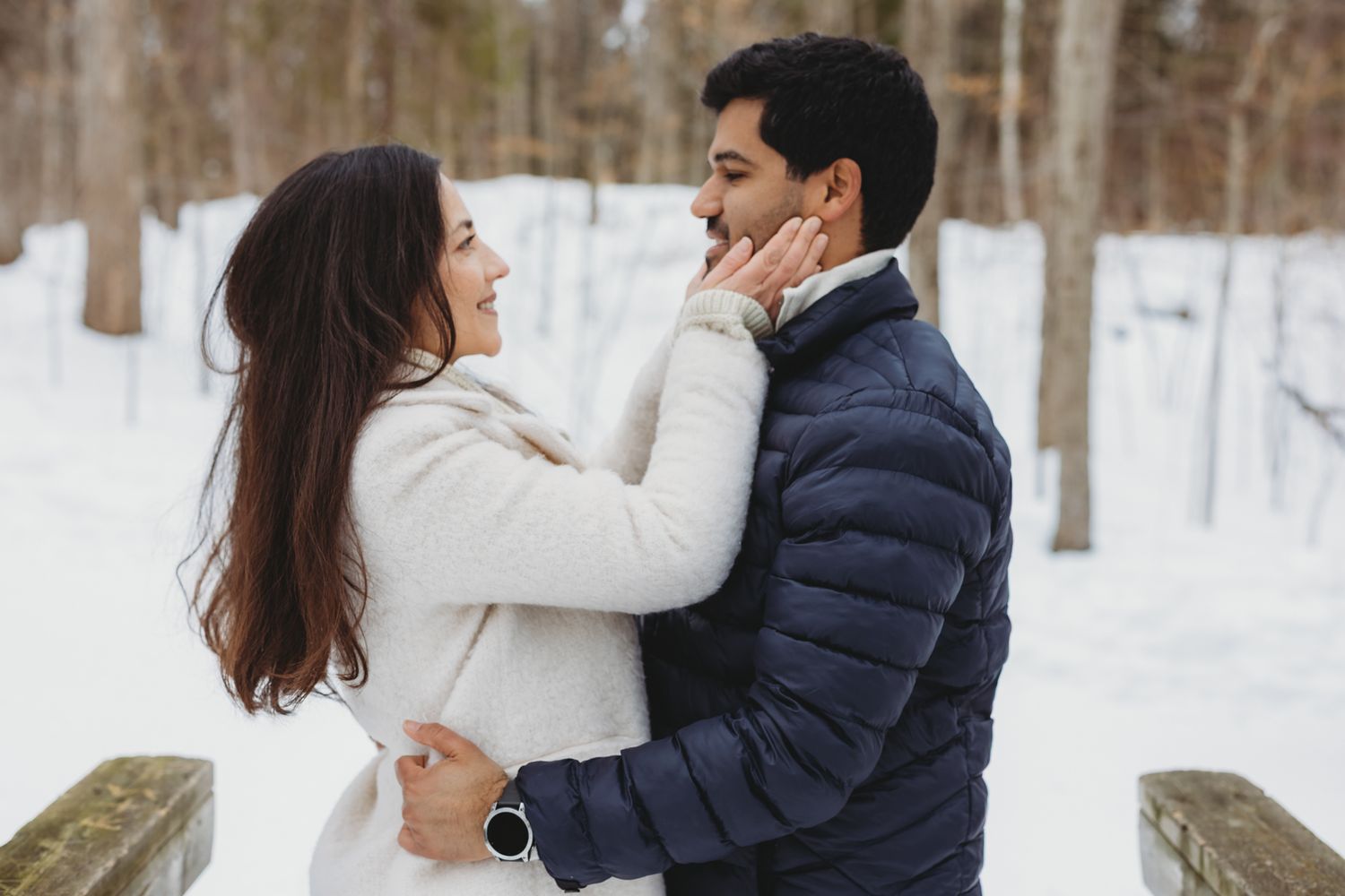 Couple embracing and smiling in a snowy winter forest, woman in white coat touching mans face romantically.
