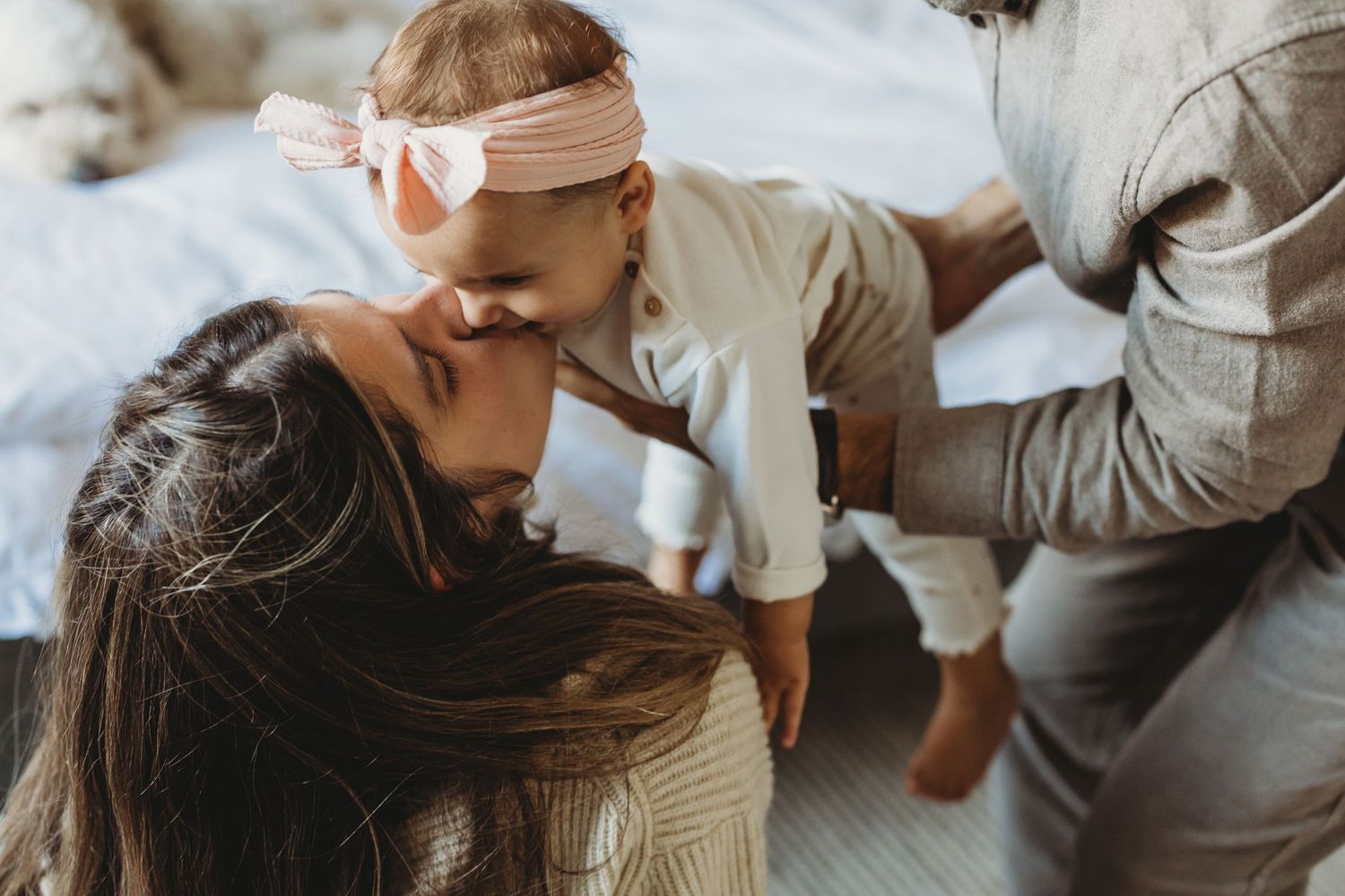 A mother lovingly kisses her baby girl wearing a pink bow headband while a partner holds the infant on a bed. Family moments.
