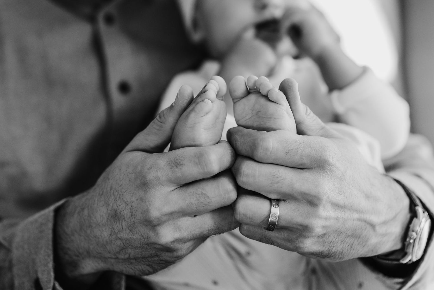 Close-up black and white photo of a father holding his newborn baby's tiny feet with wedding ring visible.