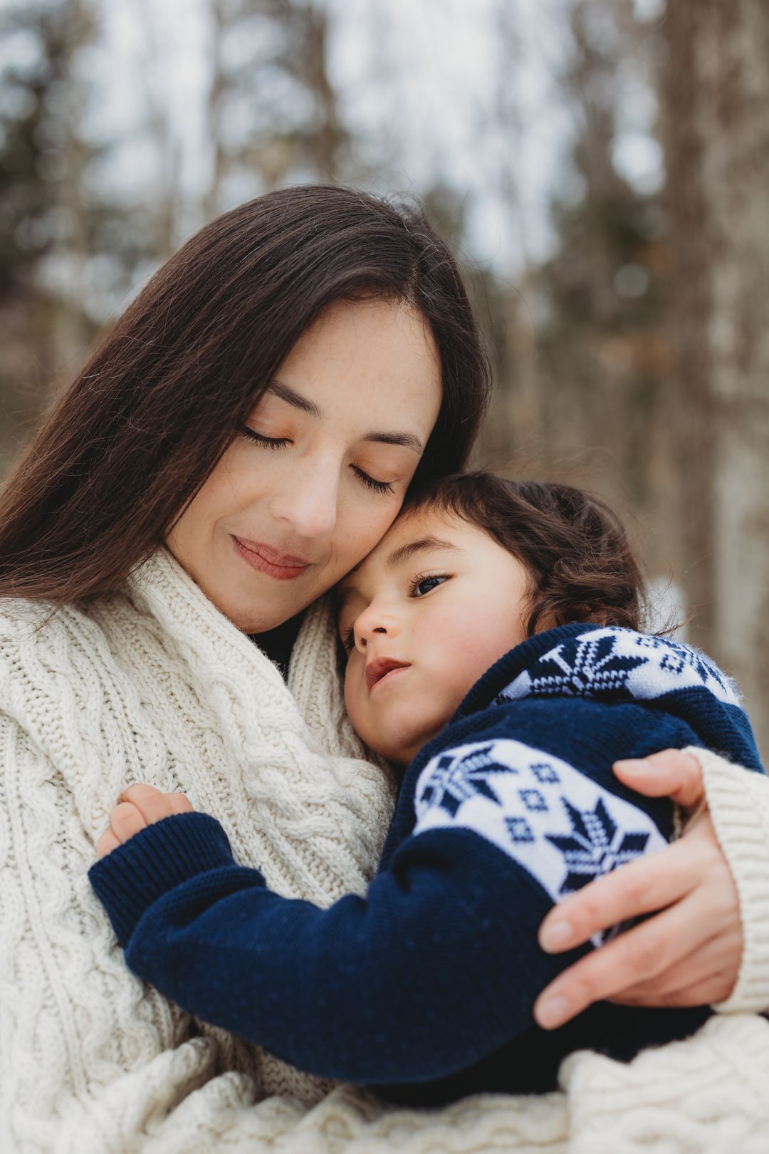 A tender moment as a dark-haired mother in a white knit sweater embraces her young child outdoors in winter.