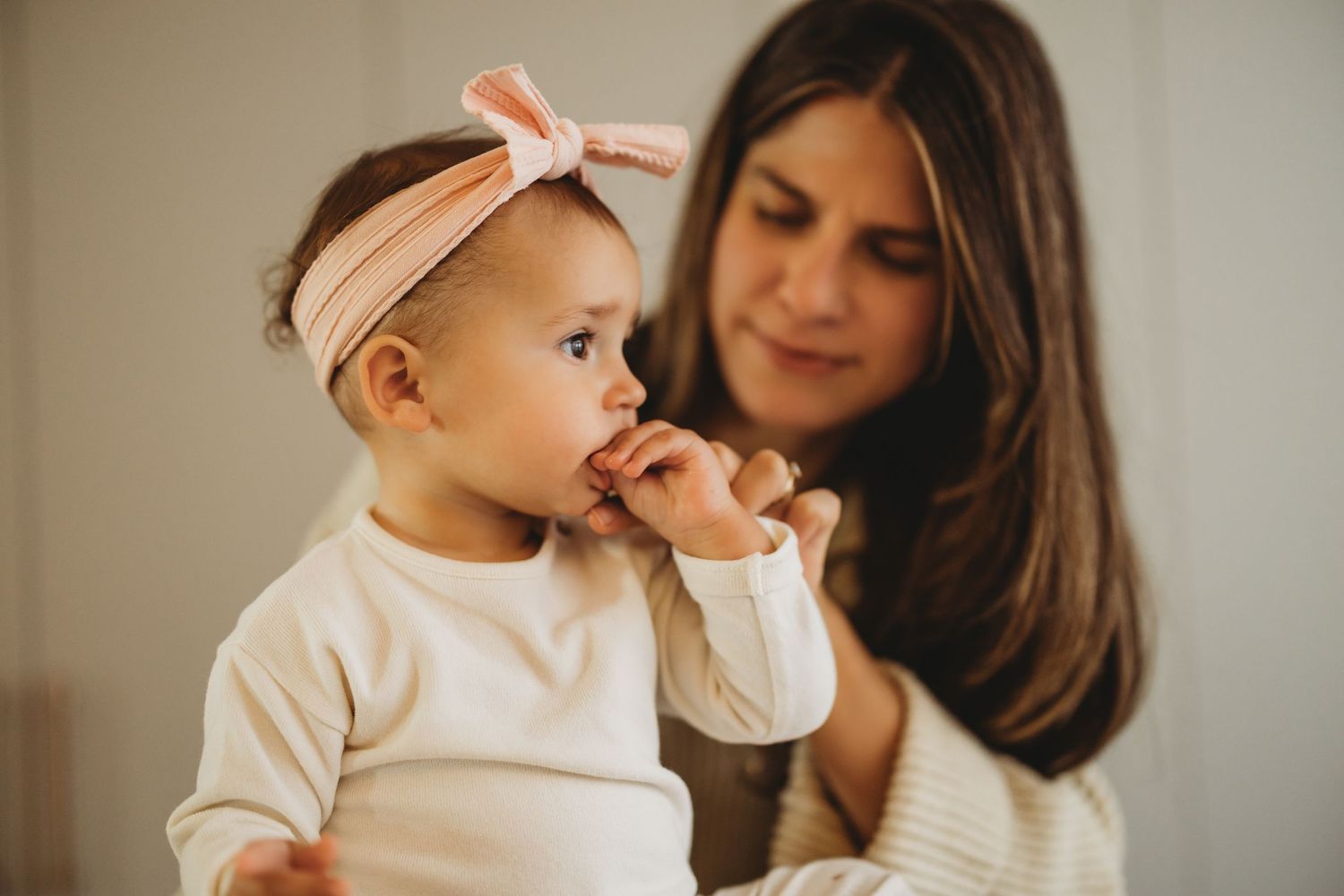 A mother tenderly holds her baby girl wearing a pink bow headband, as the infant chews on her fingers.