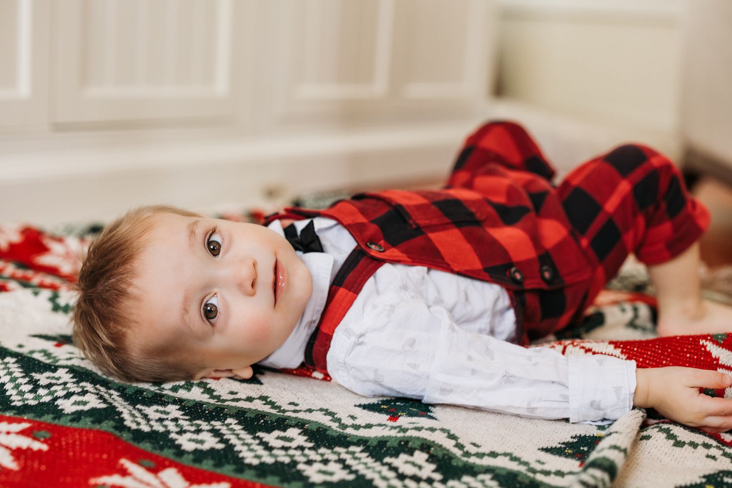 Baby in red plaid pajamas lying on a festive Christmas blanket with holiday patterns.