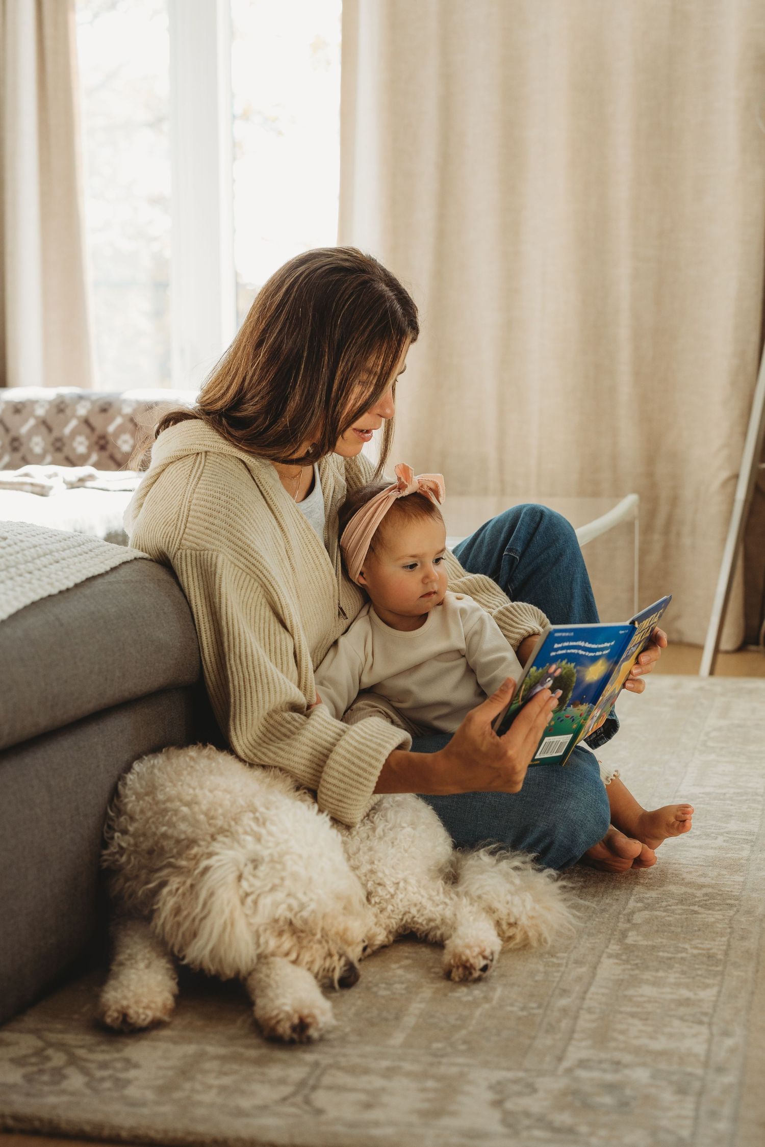 Mother reading a colorful book to her baby girl beside a fluffy white dog on a cozy living room floor.
