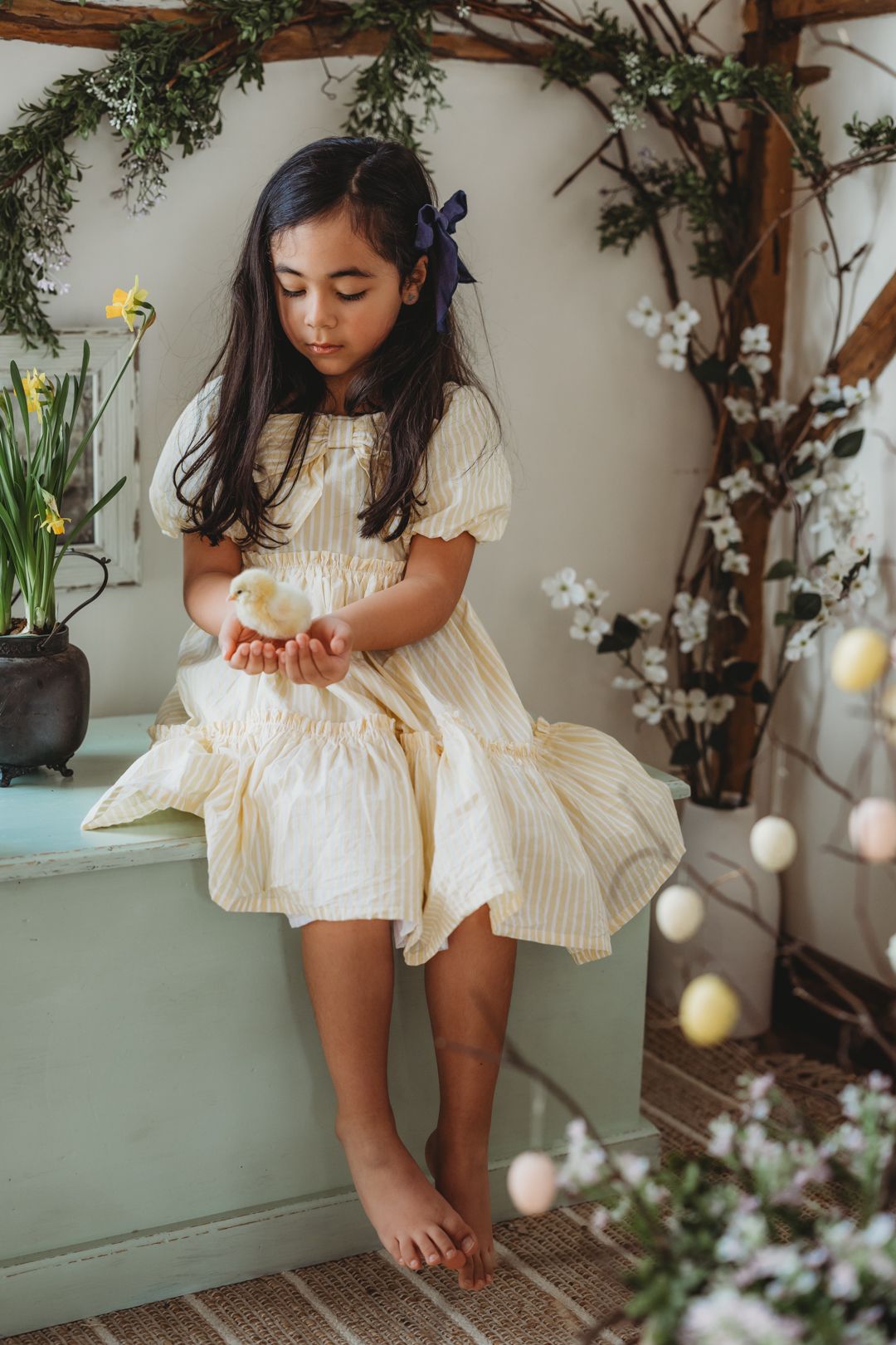 Young girl in yellow striped dress holds a baby chick while sitting in a spring Easter-themed room.
