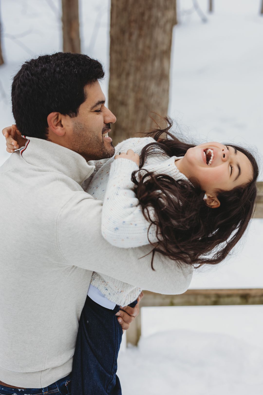 A joyful man lifts a laughing daughter in a snowy winter setting, both sharing a playful and loving moment.