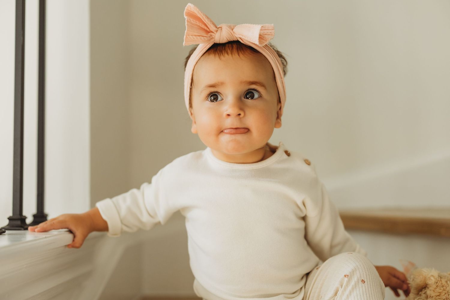 Adorable baby girl in white outfit and pink bow headband sitting on stairs with bright blue eyes.