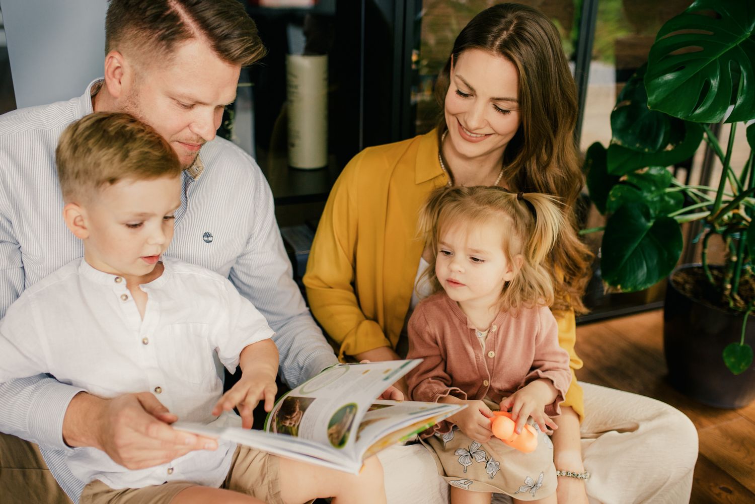 Family of four reading a colorful book together, parents smiling at children in bright home setting.