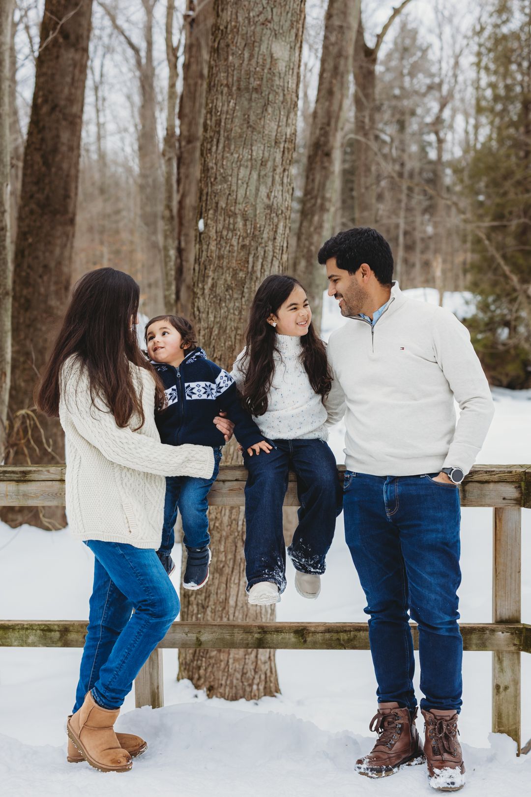 Joyful family of four enjoying winter day outdoors, children sitting on wooden fence surrounded by snow.