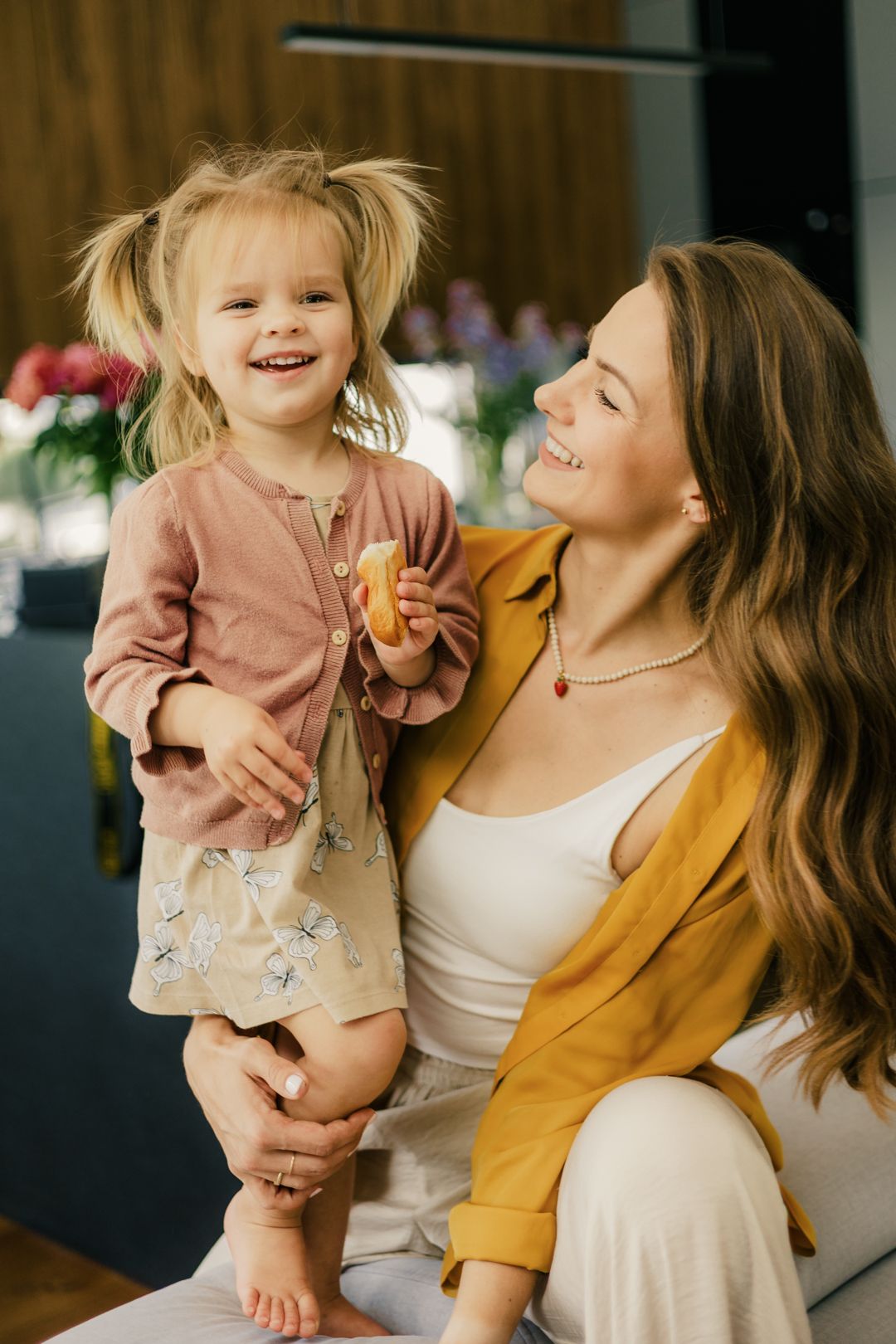 A joyful mother in a yellow jacket smiles at her laughing toddler daughter with pigtails in a modern home.