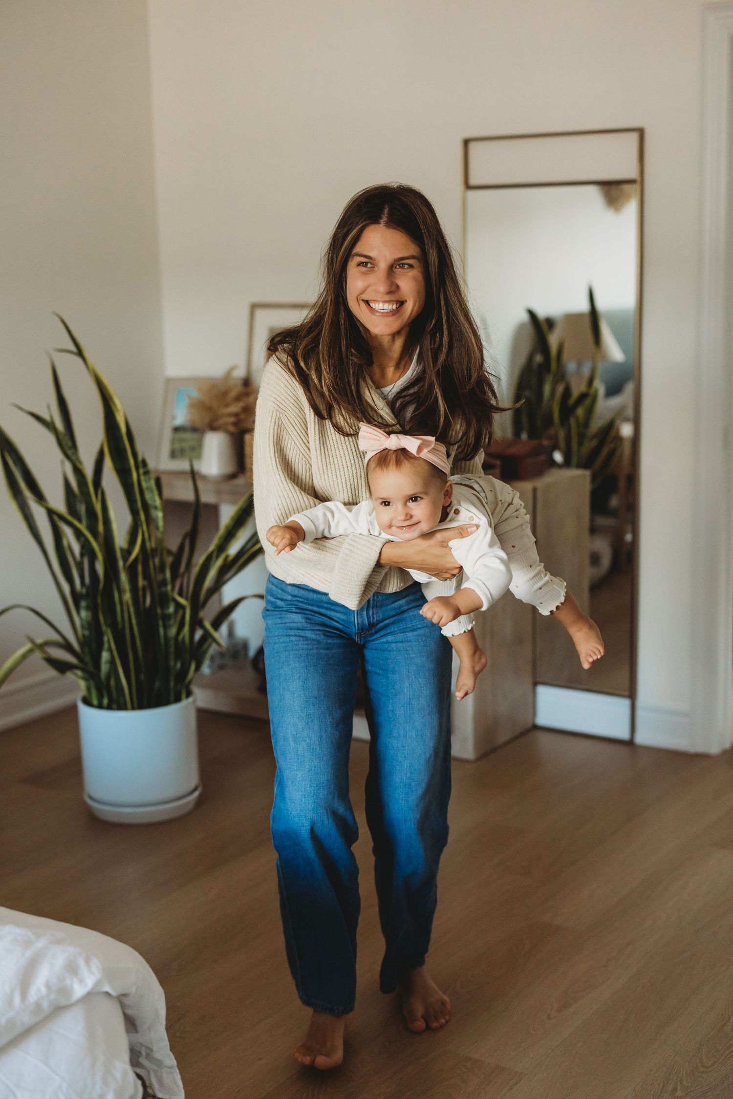 Smiling woman in jeans and cream sweater holding a baby girl with pink bow headband in a bright home.
