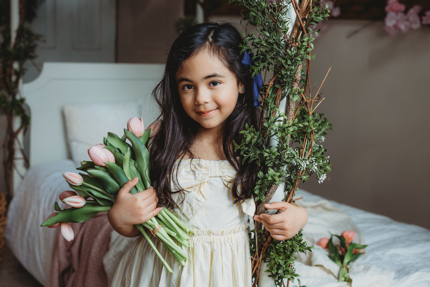 Smiling young girl in yellow dress holding pink tulips beside rustic floral branch decoration.