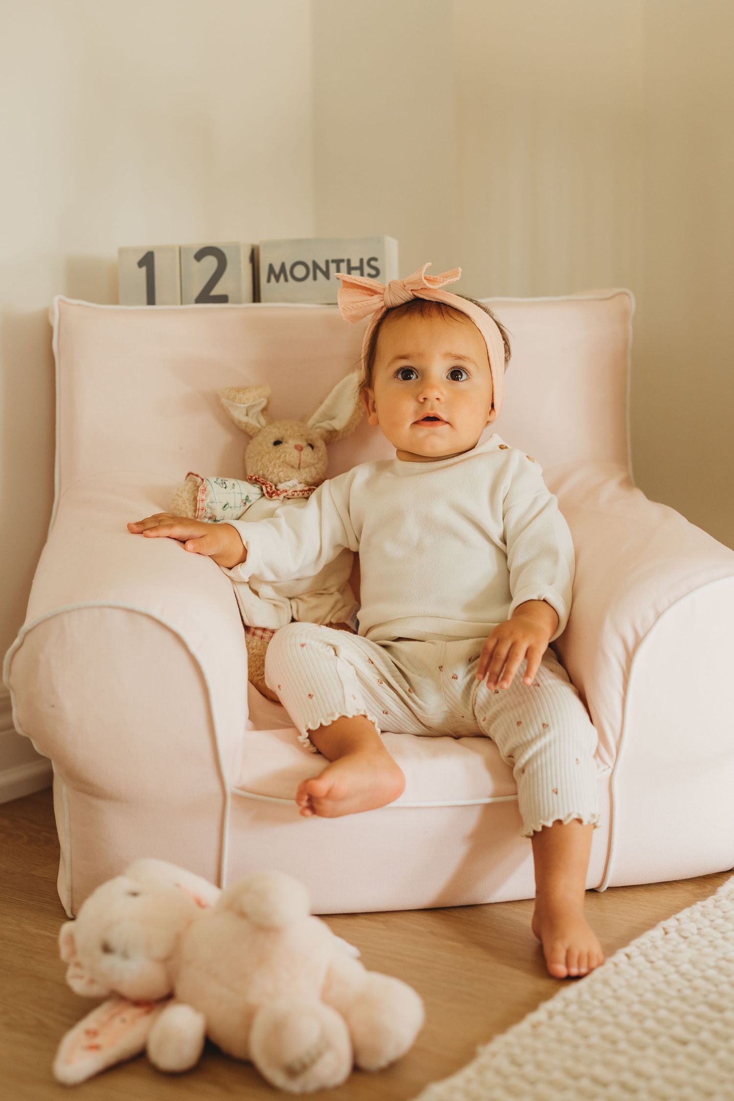 A baby girl sitting in a pink armchair with a bunny toy, celebrating her 12-month milestone.