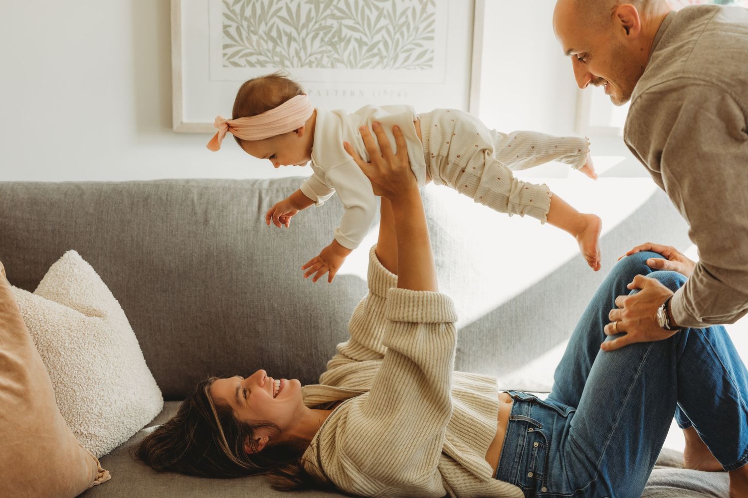 Happy family playing with baby on couch, mother lifting infant while father watches, cozy living room setting.