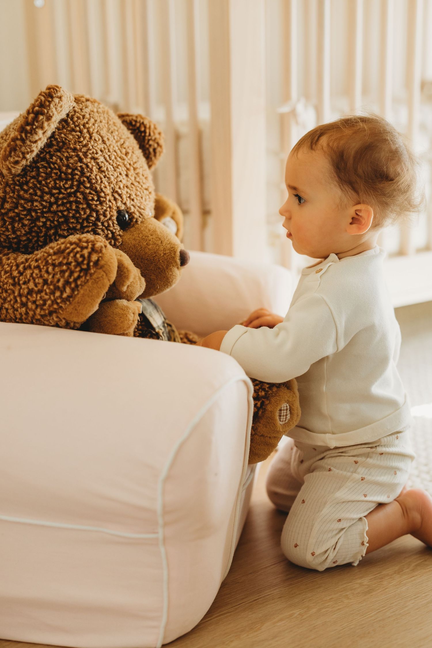 Curious baby girl in white onesie kneeling on floor interacting with a large brown teddy bear.