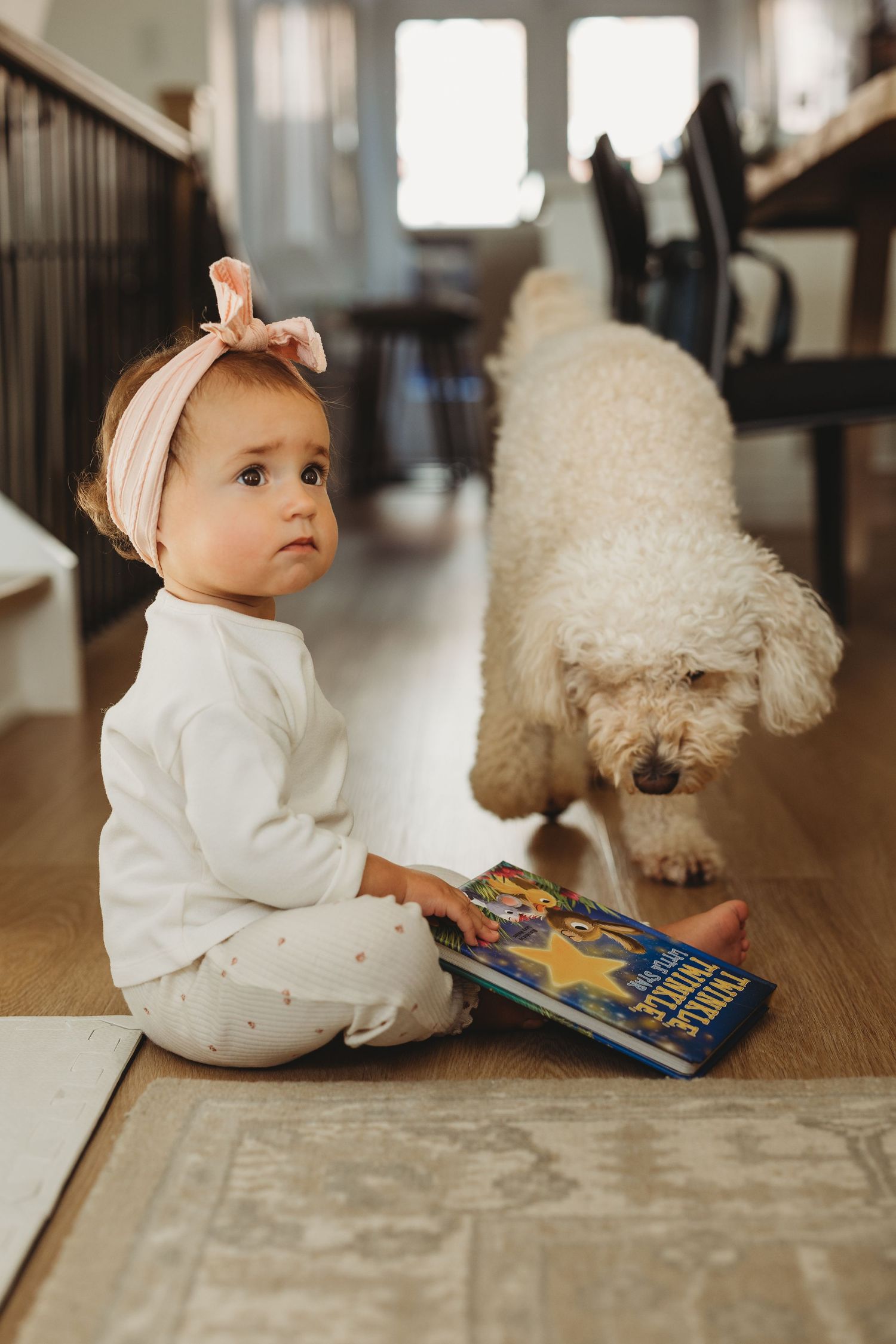 Baby girl with pink bow sits on floor holding a book while a fluffy white dog sniffs nearby.