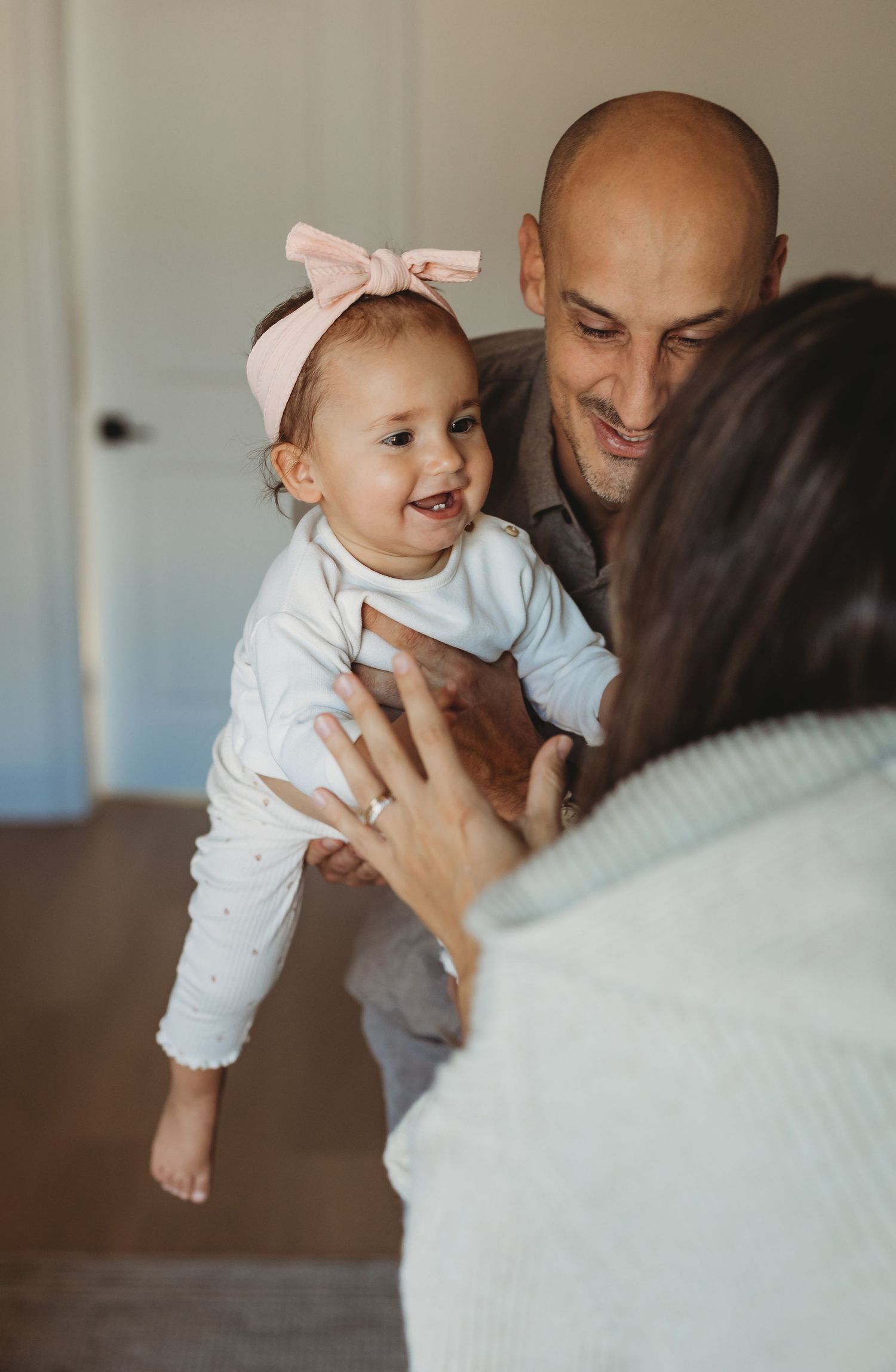 A bald father holds a smiling baby girl with a pink bow headband while mother reaches out indoors.