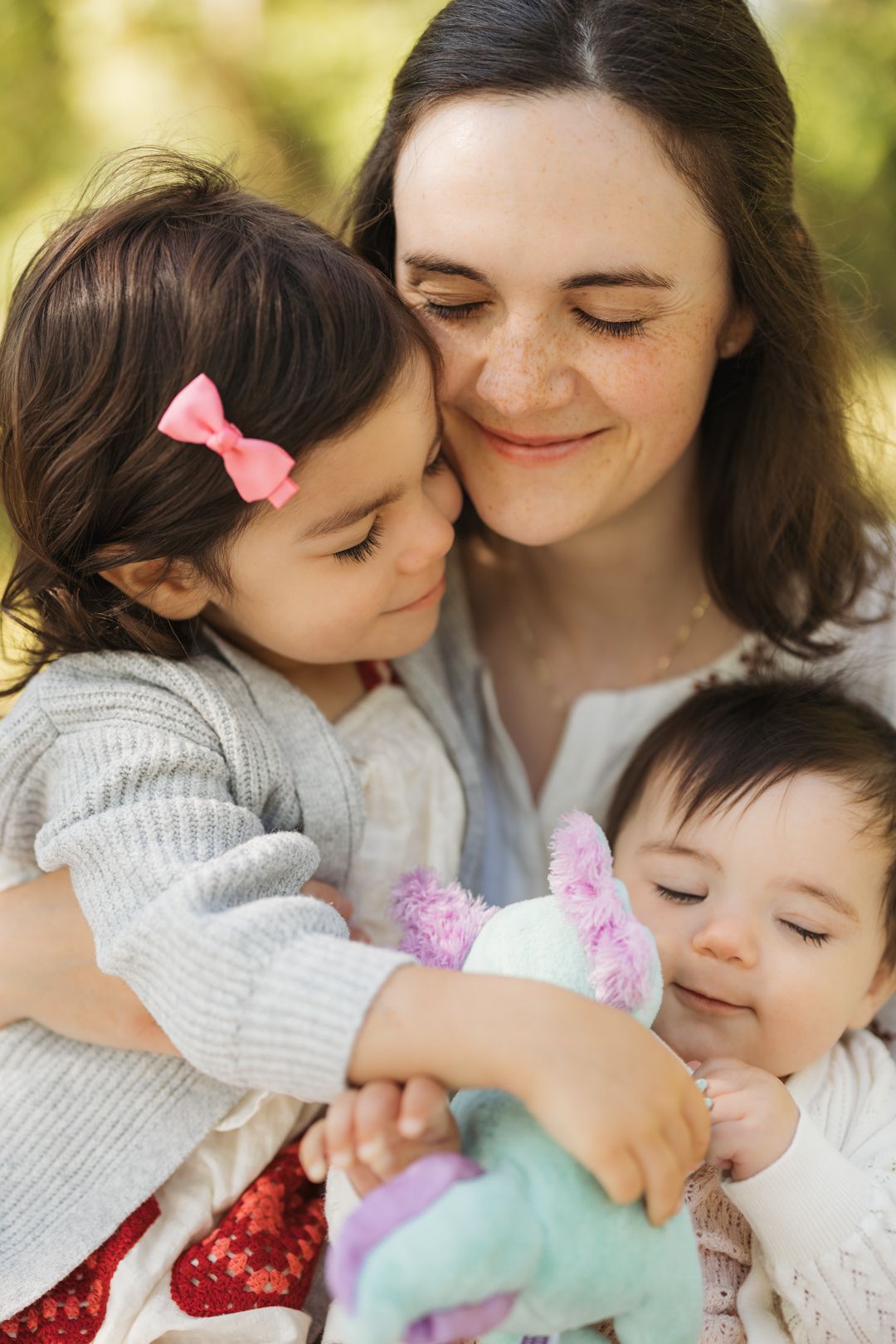 A mother tenderly embraces her two young daughters outdoors, one toddler with a pink bow hugging her cheek. Family moments in Burlington, ON