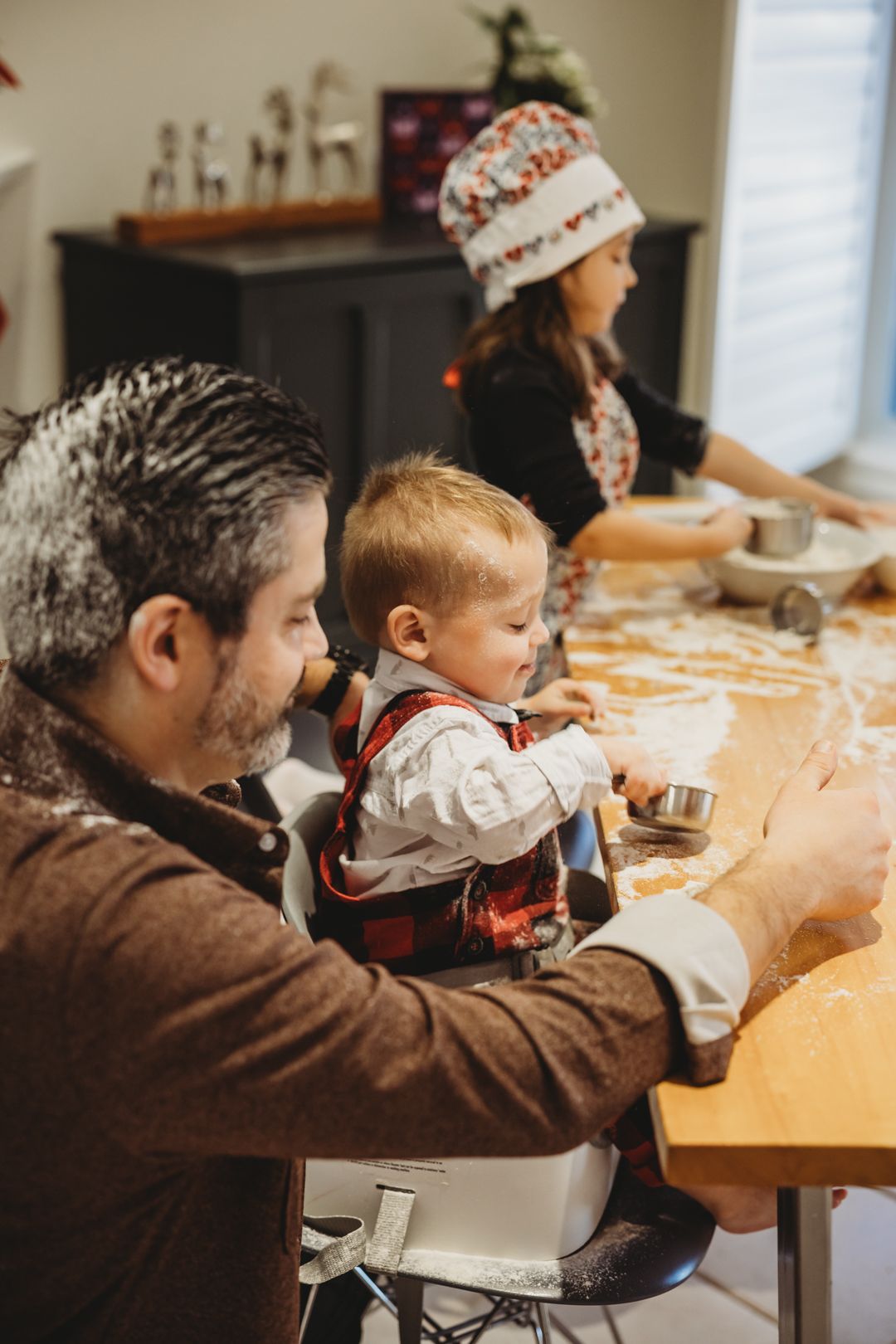 A family bakes holiday cookies together at a wooden table, with a father holding a toddler and a woman wearing a festive hat.