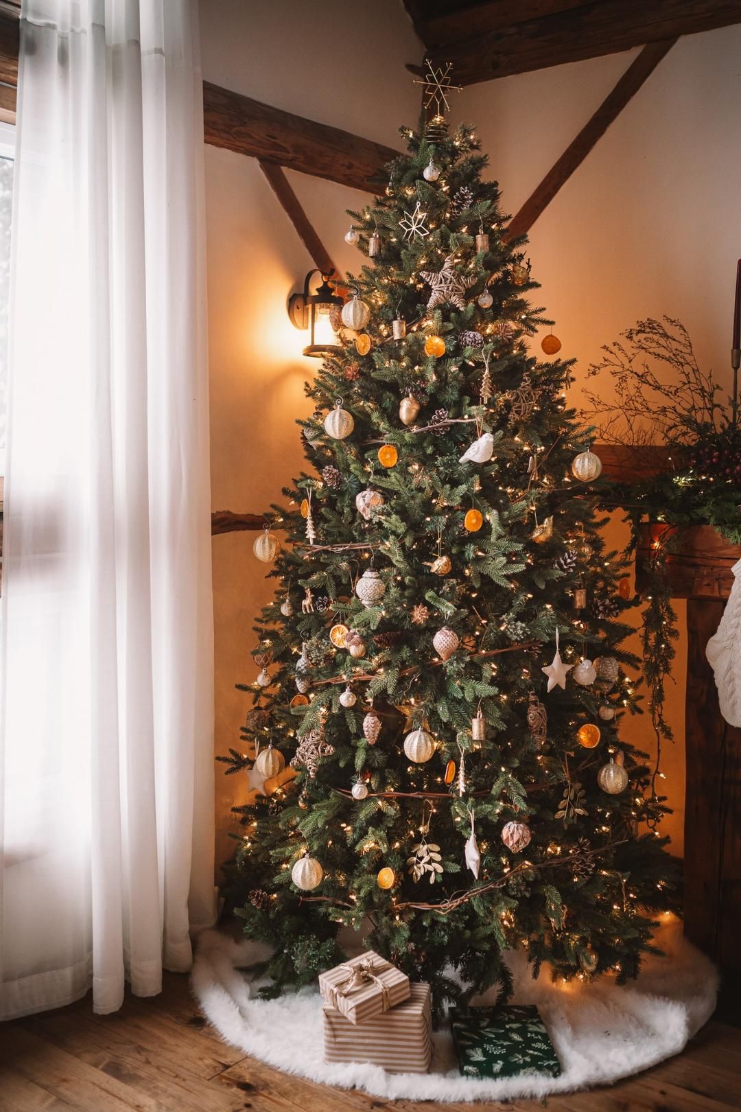 Decorated Christmas tree with gold ornaments, dried oranges, and fairy lights, with gifts underneath in a cozy rustic room.