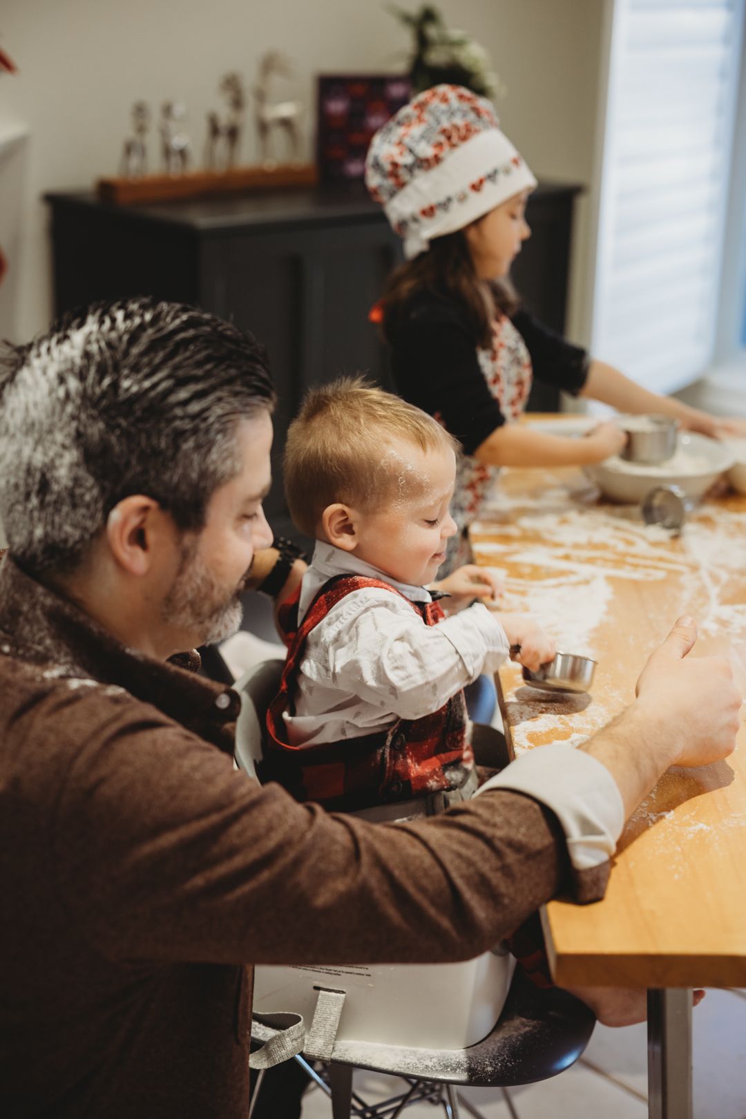 Family baking holiday cookies together at kitchen table, father holding toddler while woman in festive hat works with dough.
