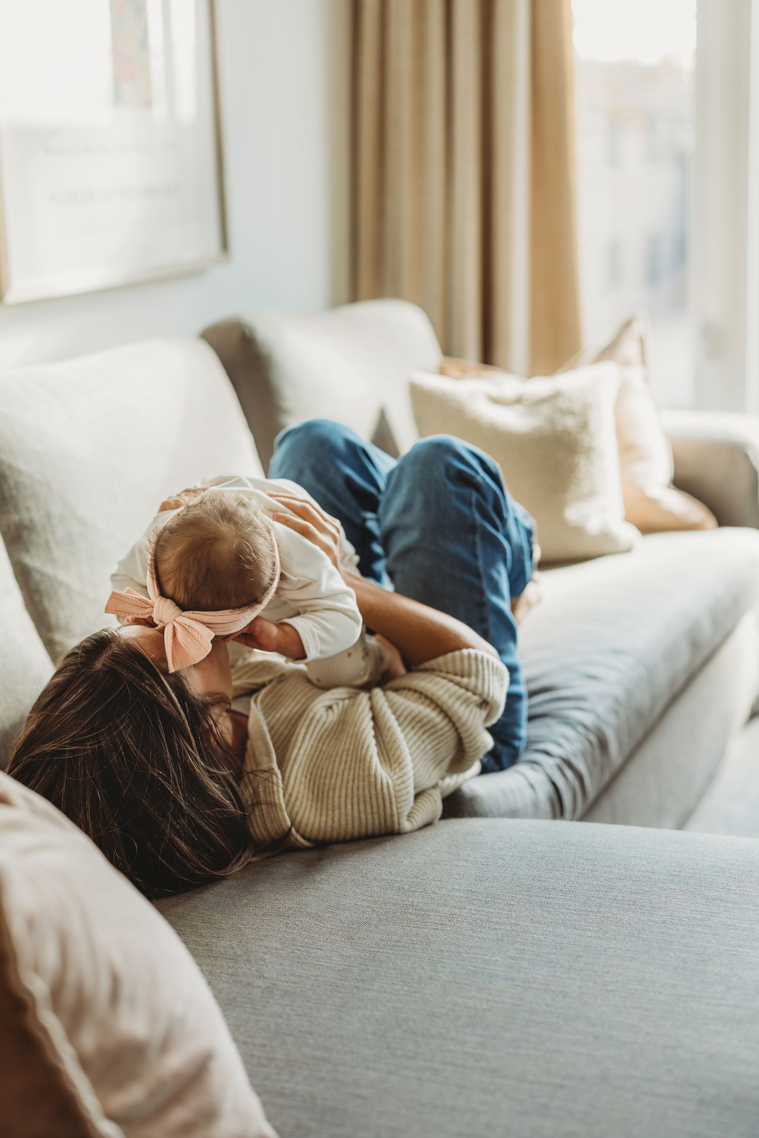 Mother lying on couch cuddling newborn baby wearing pink bow, cozy living room setting with natural light.