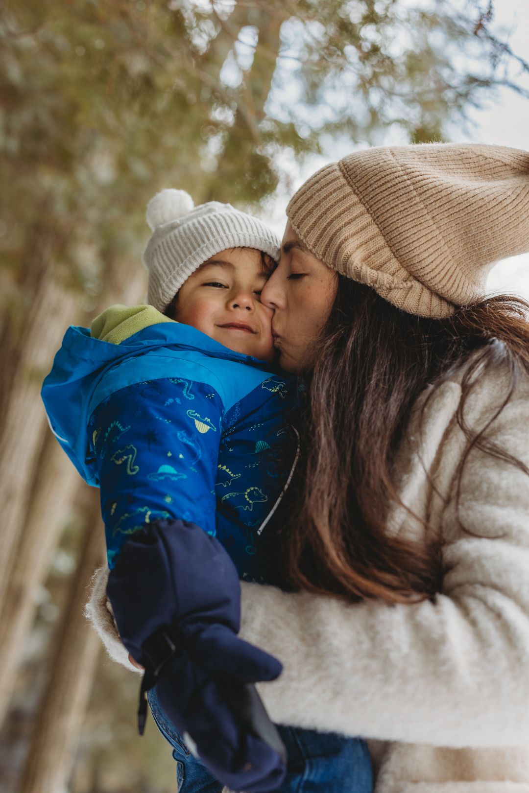 Mother in beige hat kisses toddler in blue winter jacket outdoors in snowy forest.