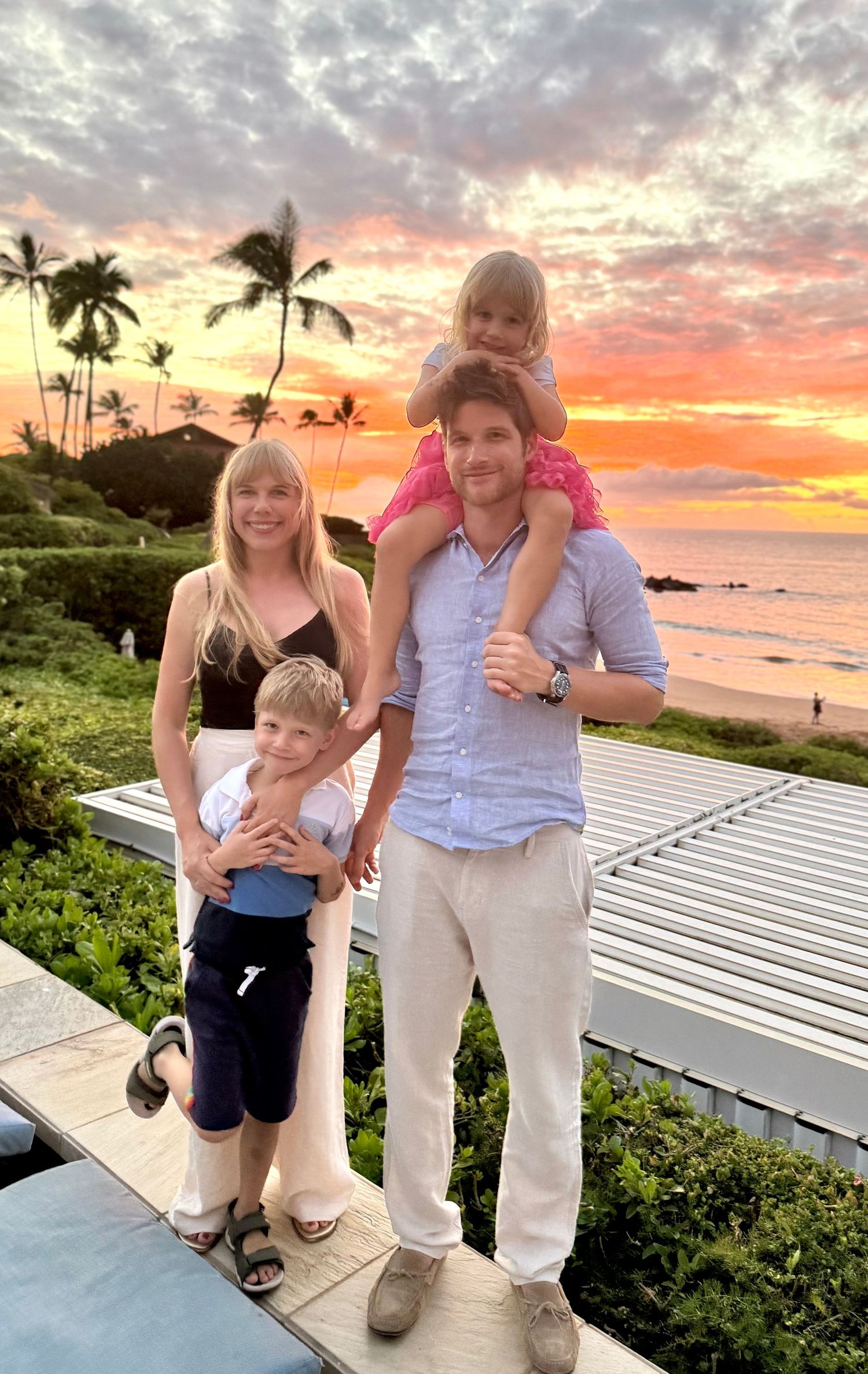 Family of four posing at sunset on a tropical beach with palm trees and vibrant orange sky.