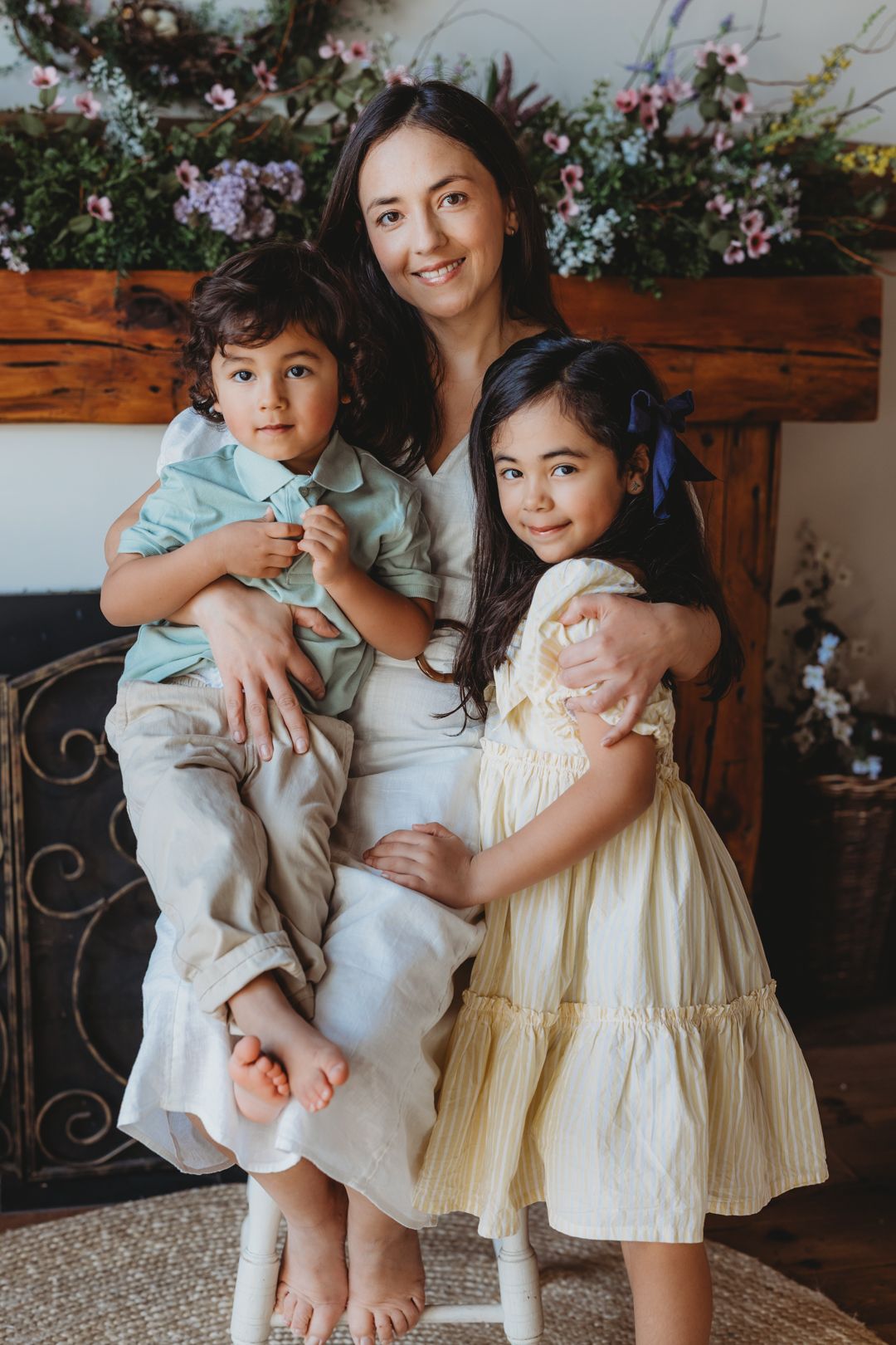 Loving mother sitting with two young children in front of floral fireplace mantel backdrop.