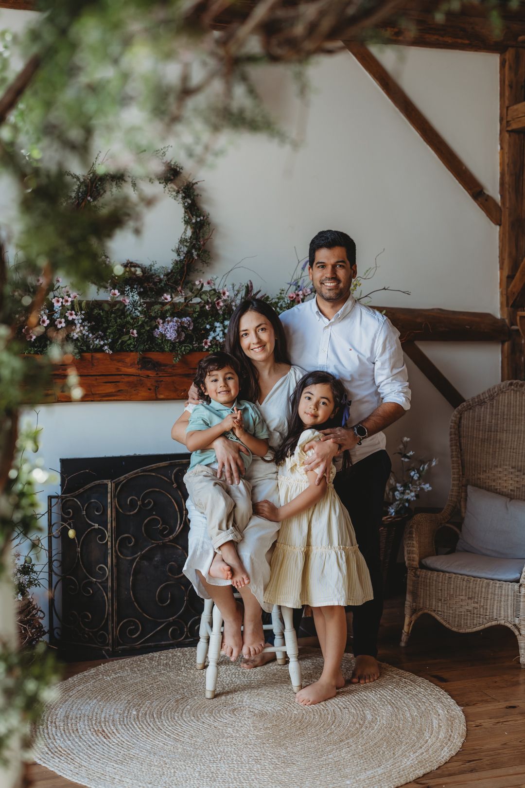 Smiling family of four poses in a rustic room decorated with greenery, flowers, and a fireplace.